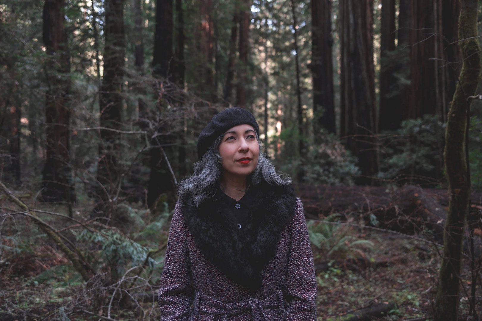 A woman with gray hair wearing a black beret and a fur-collared coat stands in a forest among tall trees.