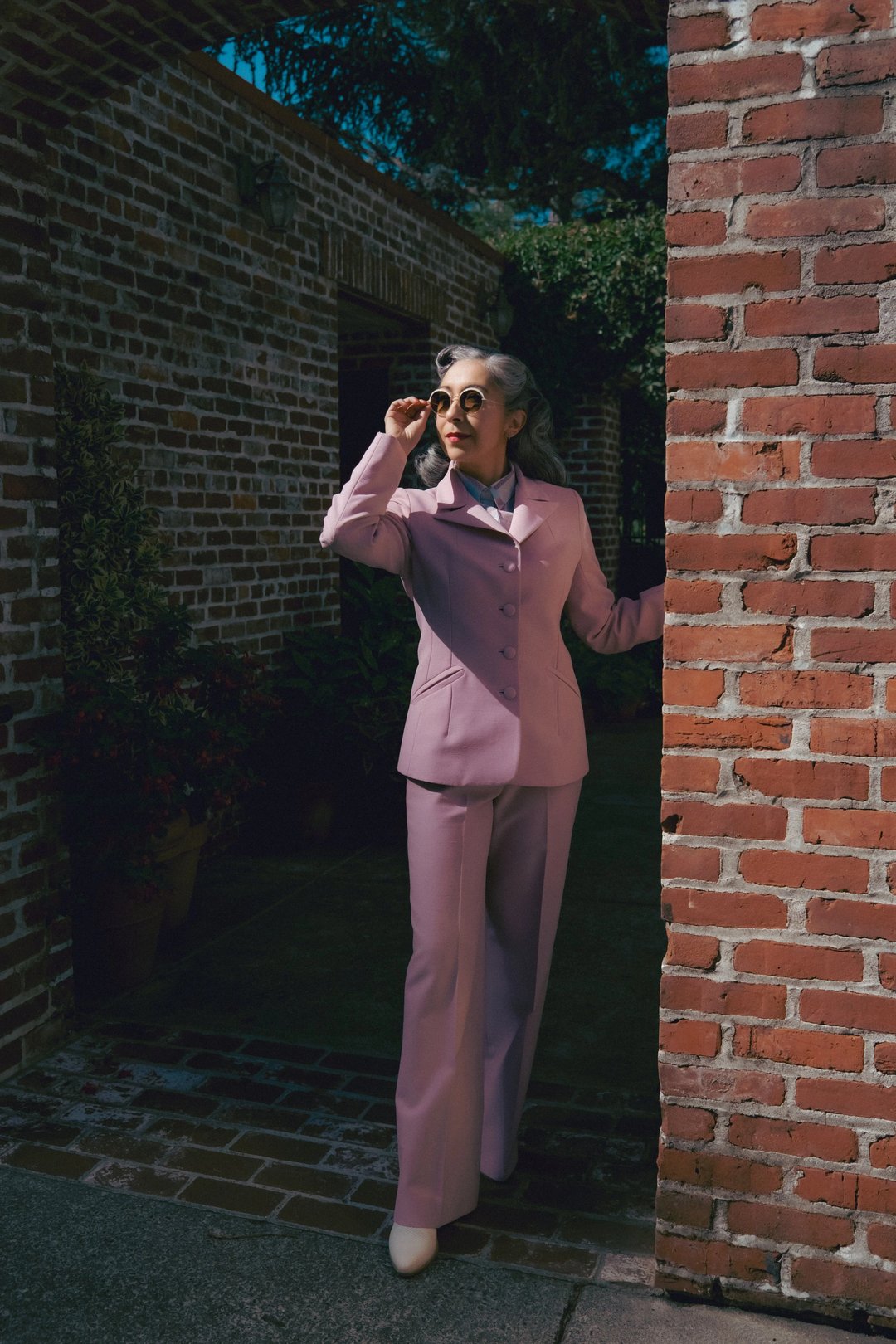 A woman in a pastel purple suit and sunglasses poses beside a brick wall, exuding a stylish and confident demeanor.