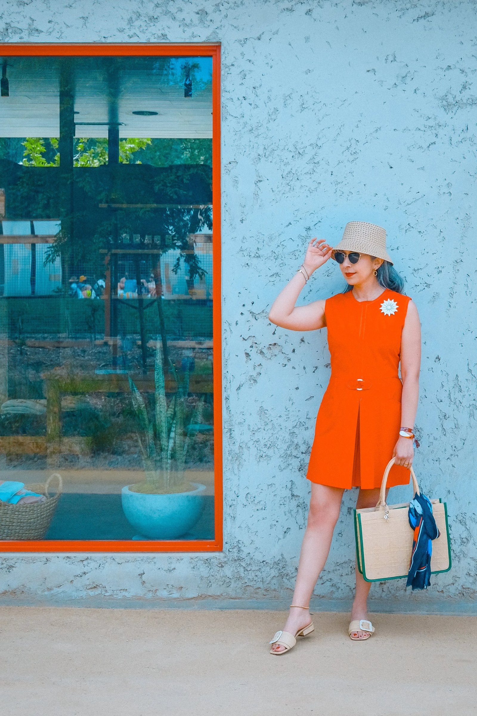 A woman in an orange dress stands beside a blue wall with a large red-framed window, wearing a hat and sunglasses, and holding a handbag.