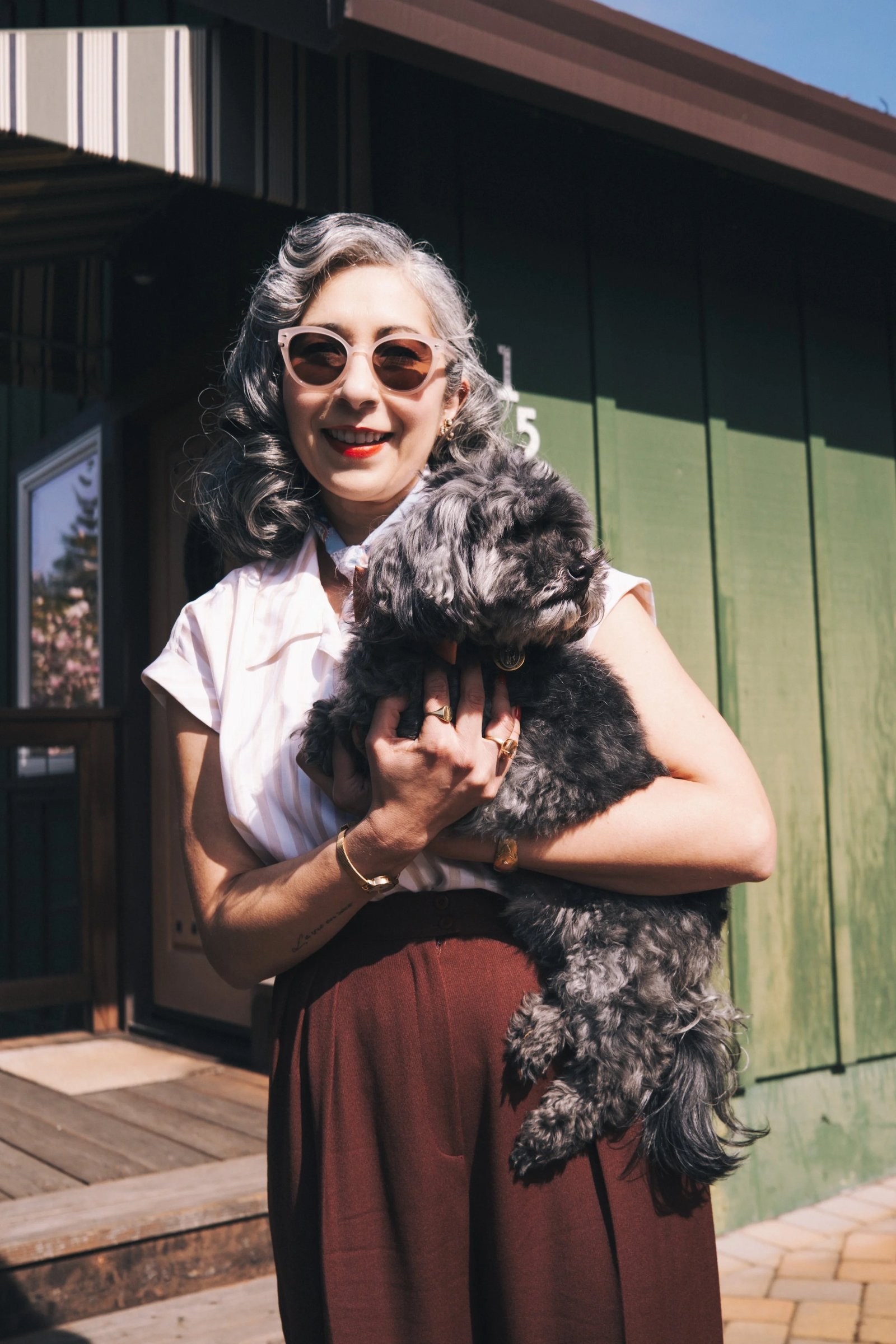 A woman with gray hair wearing sunglasses holds a small black dog, standing in front of a green building.