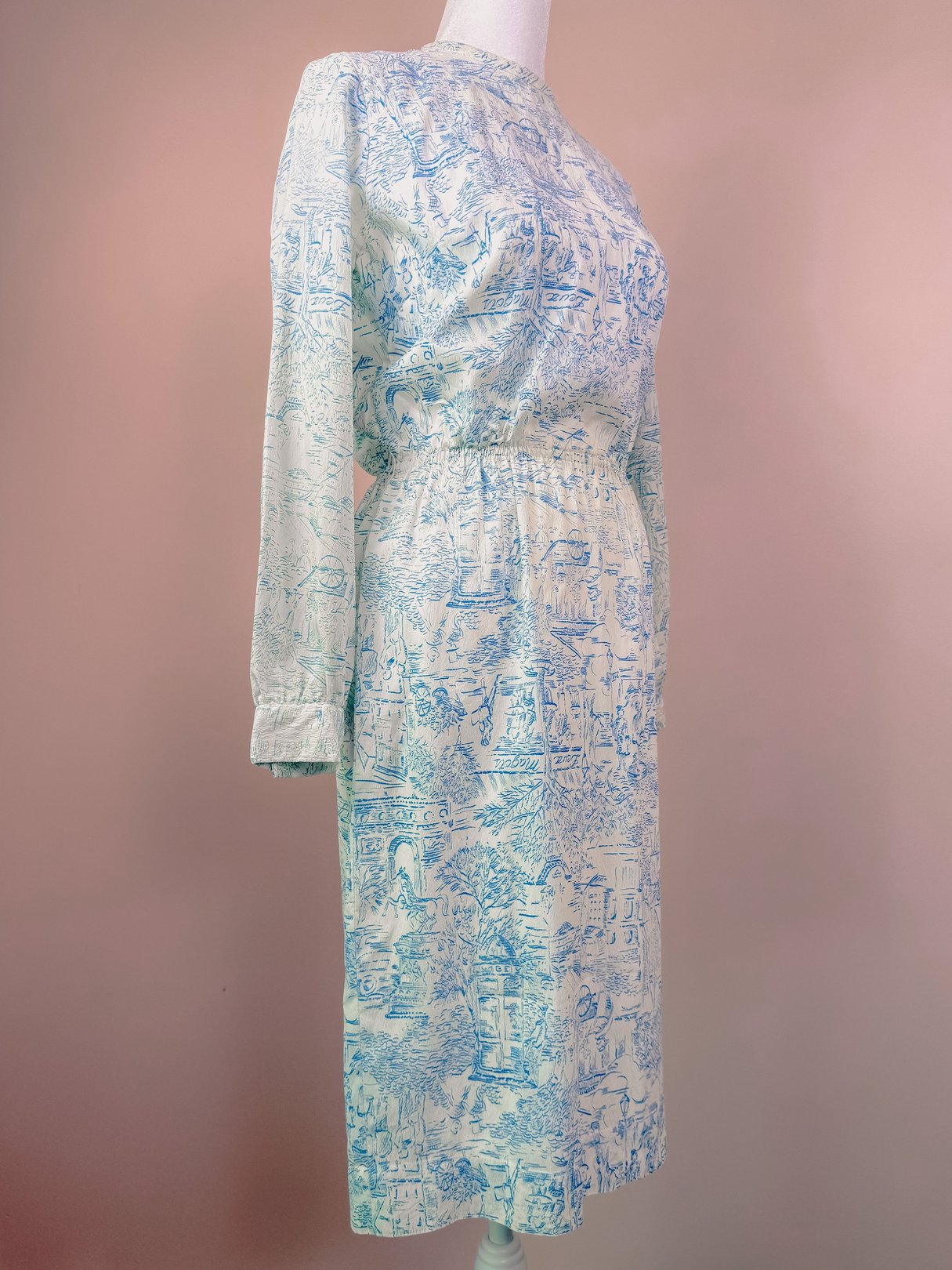 The Paris Montage Dress is a blue 1960s women's dress featuring a cinched waist and long sleeves with a scenic print.