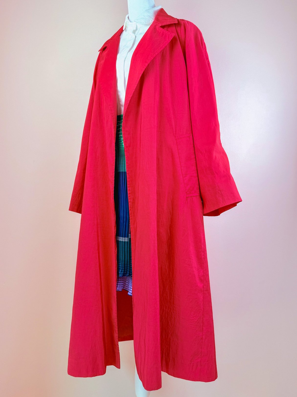 A women's red rain jacket from the 1980s, featuring a long, loose fit and classic collar, displayed on a mannequin against a pastel background.
