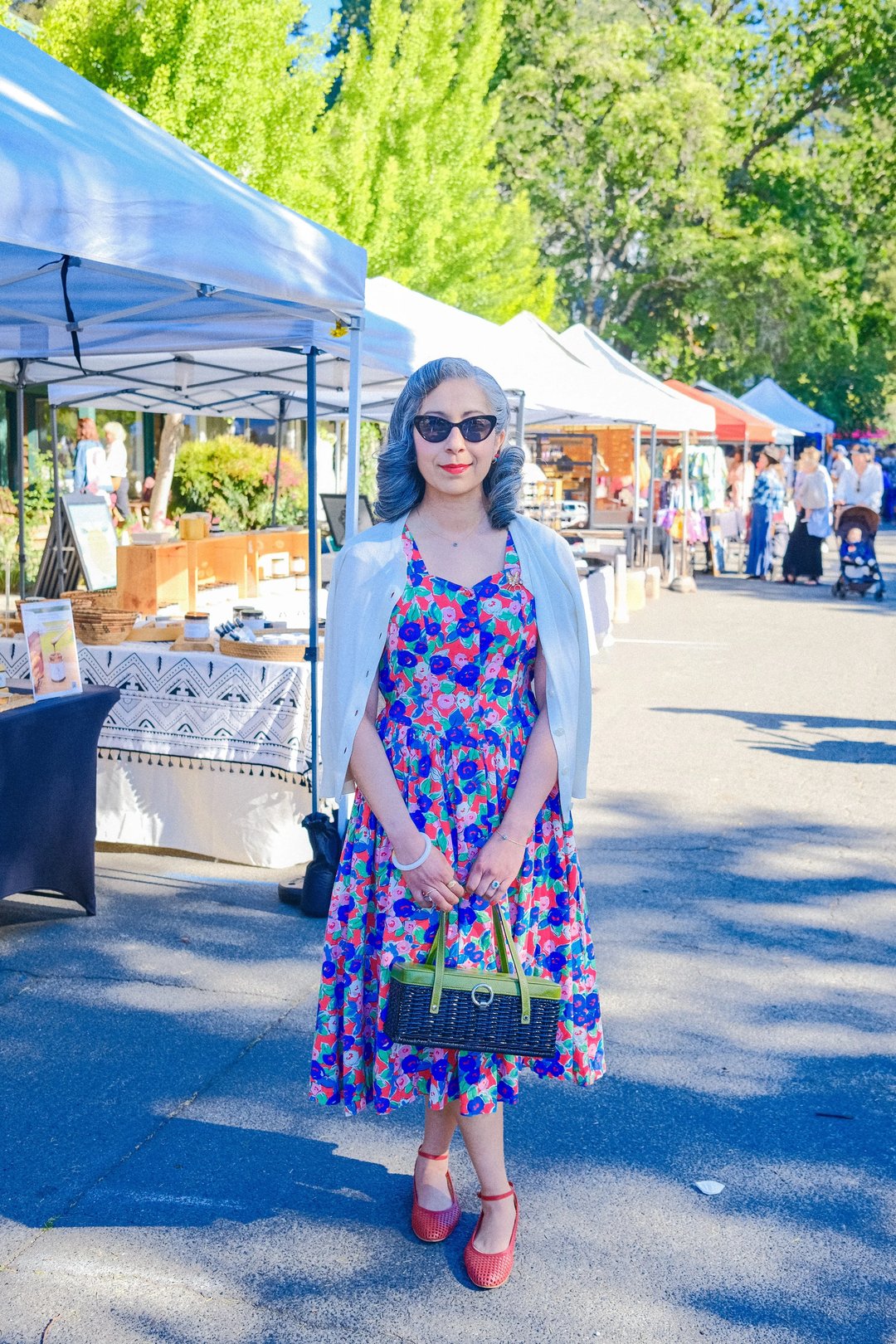A woman wearing a floral dress and sunglasses stands confidently at an outdoor market surrounded by tents and people.