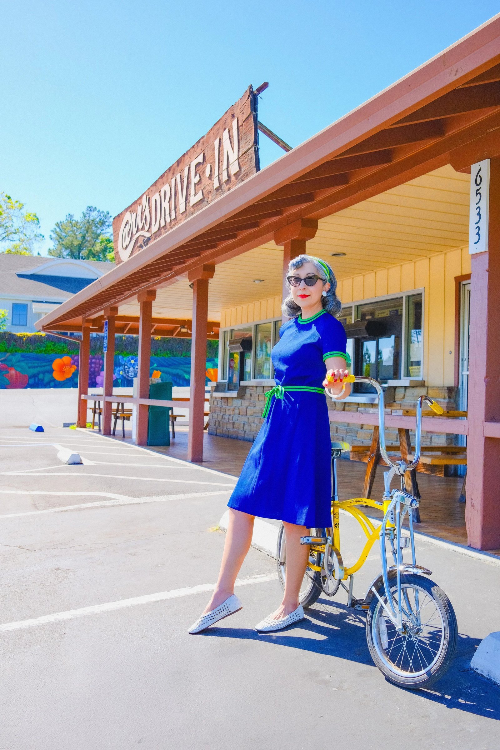 A woman in a blue dress poses with a yellow bicycle in front of a vintage-style building.