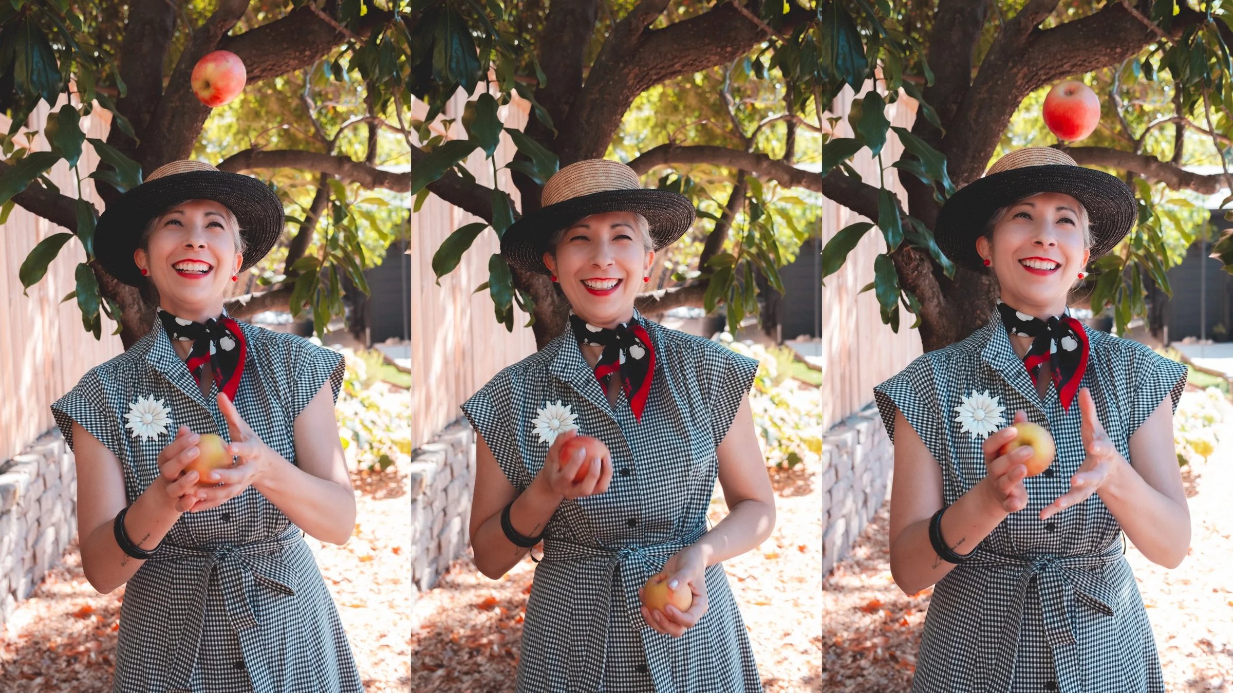 A woman wearing a straw hat and a checkered dress smiles while holding an apple in a garden setting.