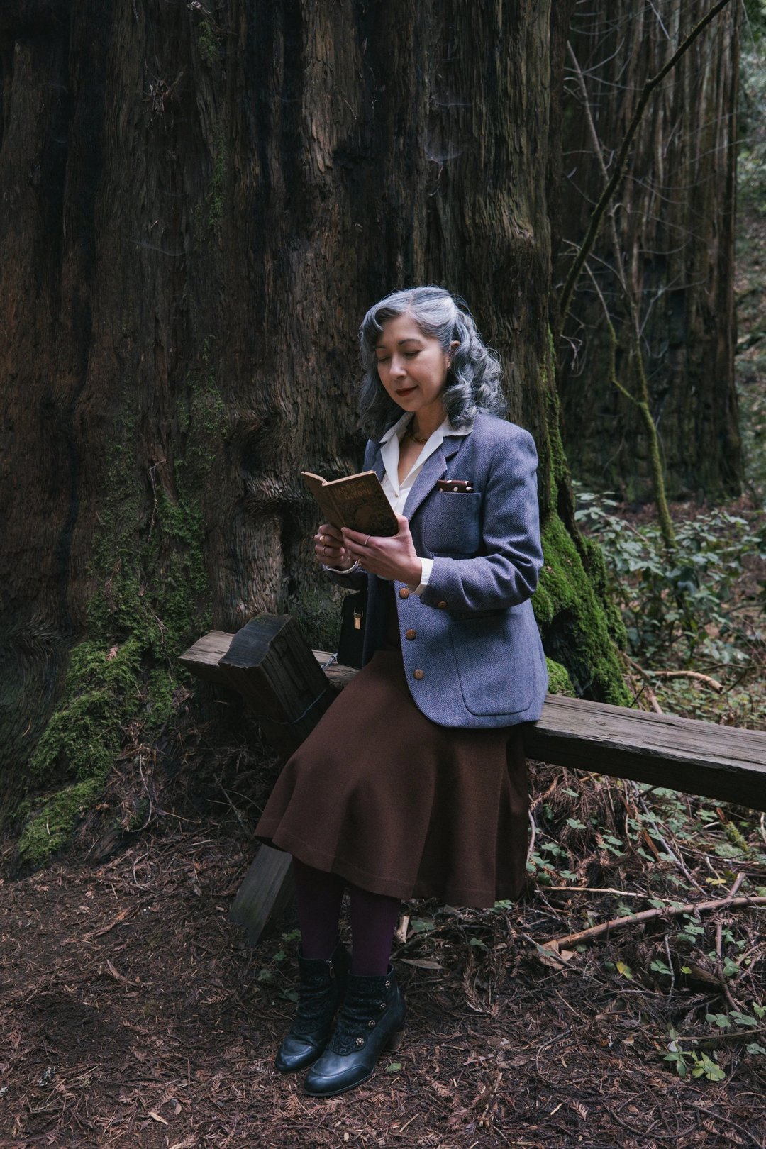 A woman in a vintage outfit sits on a wooden bench in a forest, reading a book.