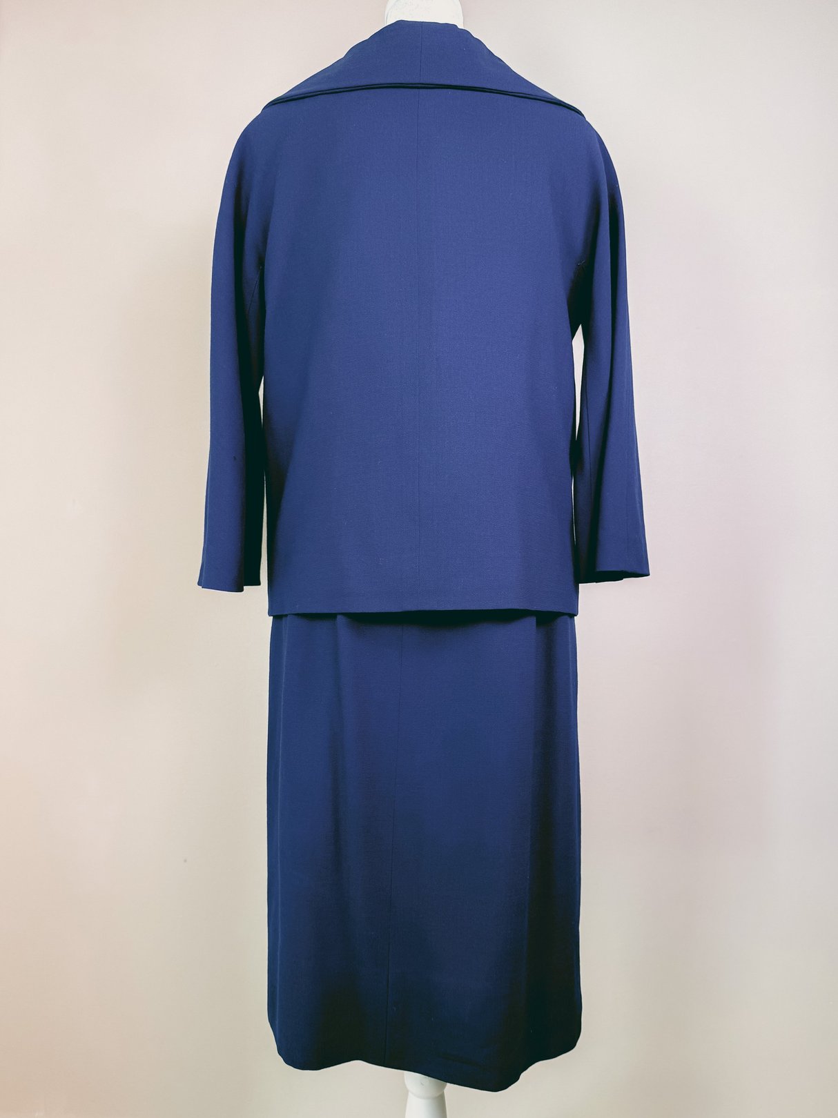 Navy blue 1960s women's dress set featuring a fitted skirt and fitted jacket with three-quarter sleeves displayed from the back.