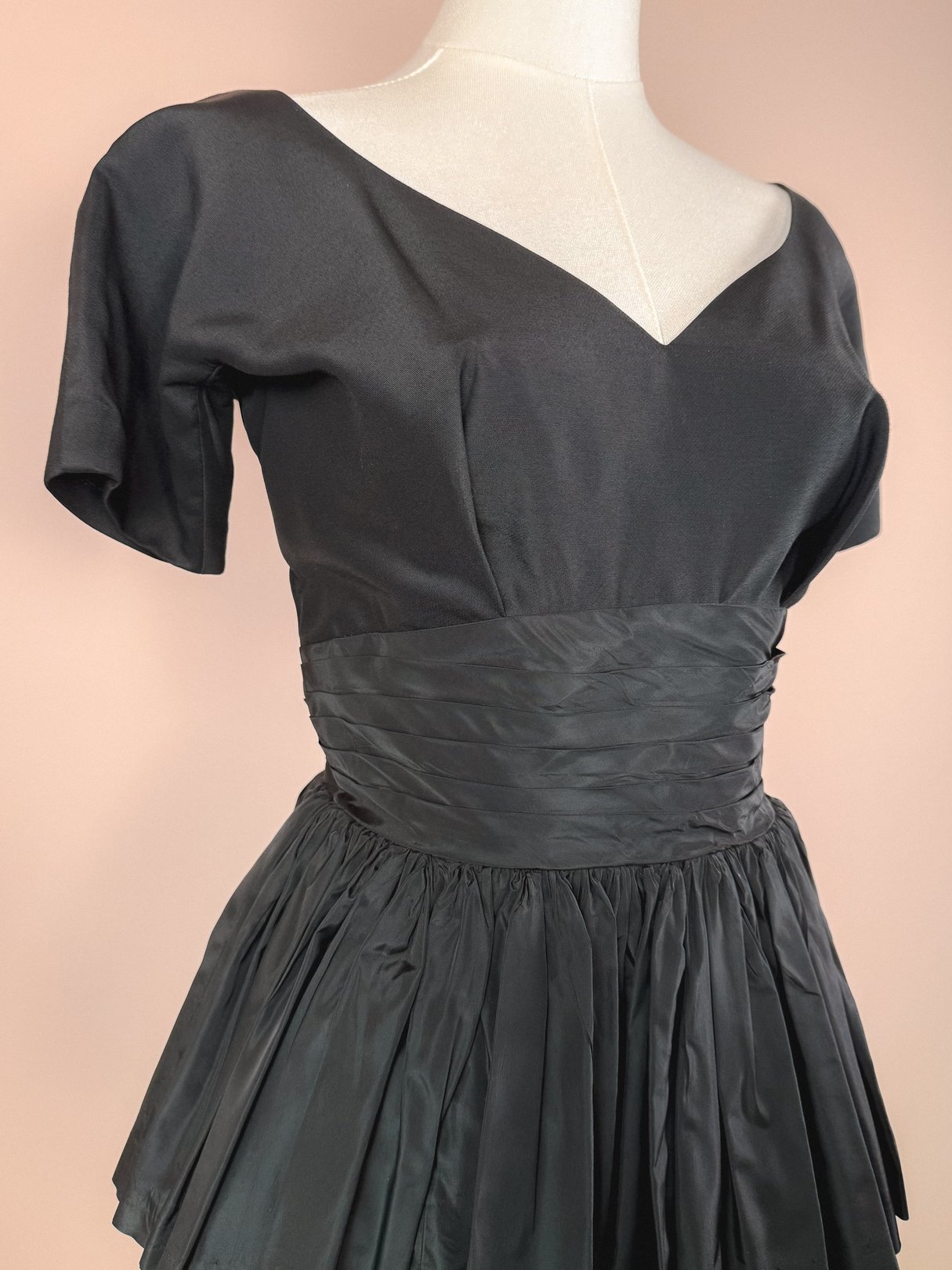 1950s black dress for women featuring a fitted bodice with a pleated waist and flared skirt.