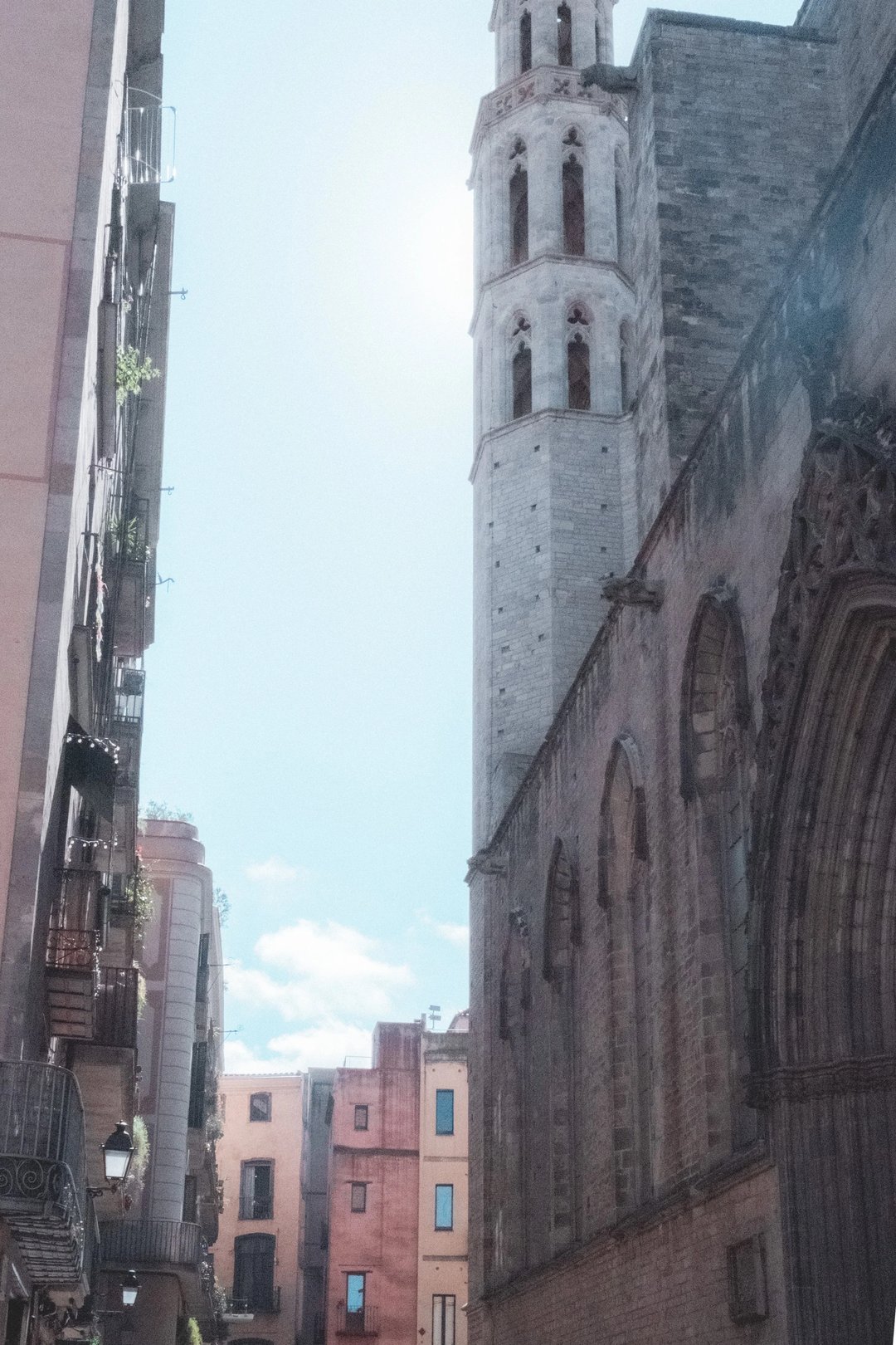 A narrow street in Barcelona featuring a historic church with a tall tower and colorful buildings.