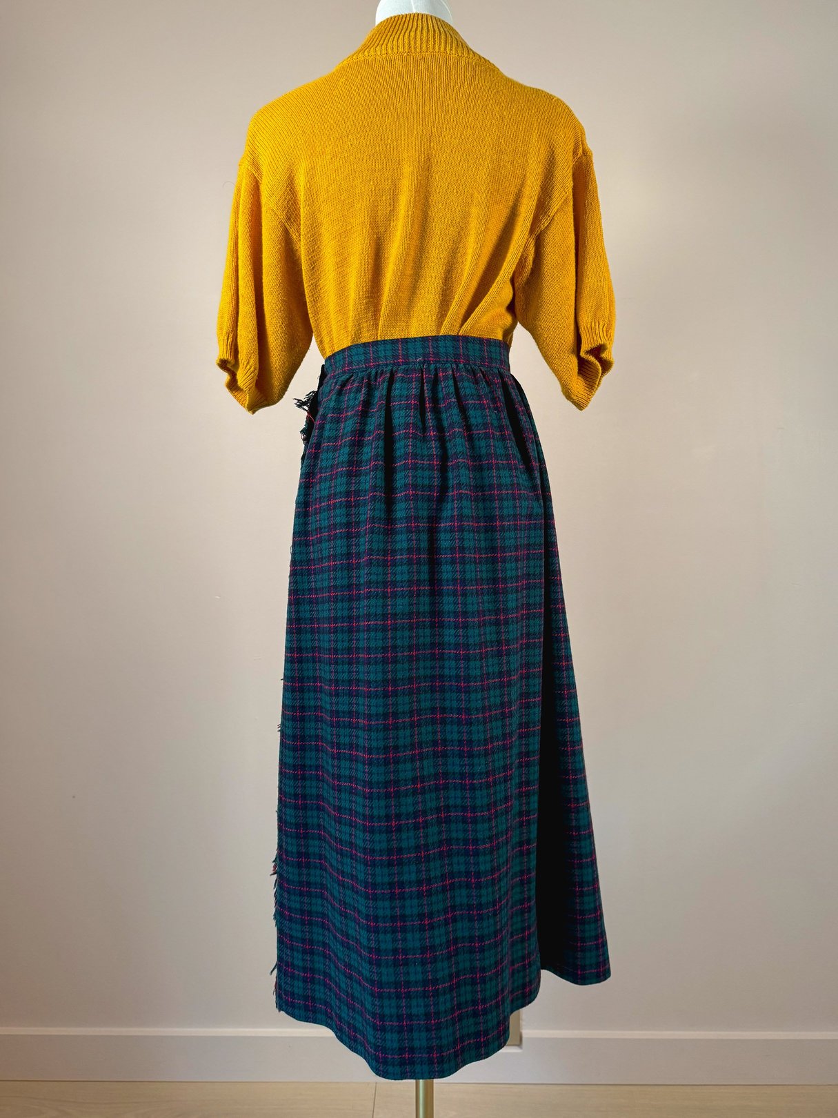 A back view of a green plaid skirt with a gathered waistband, styled with a yellow top, representing 1970s women's fashion.