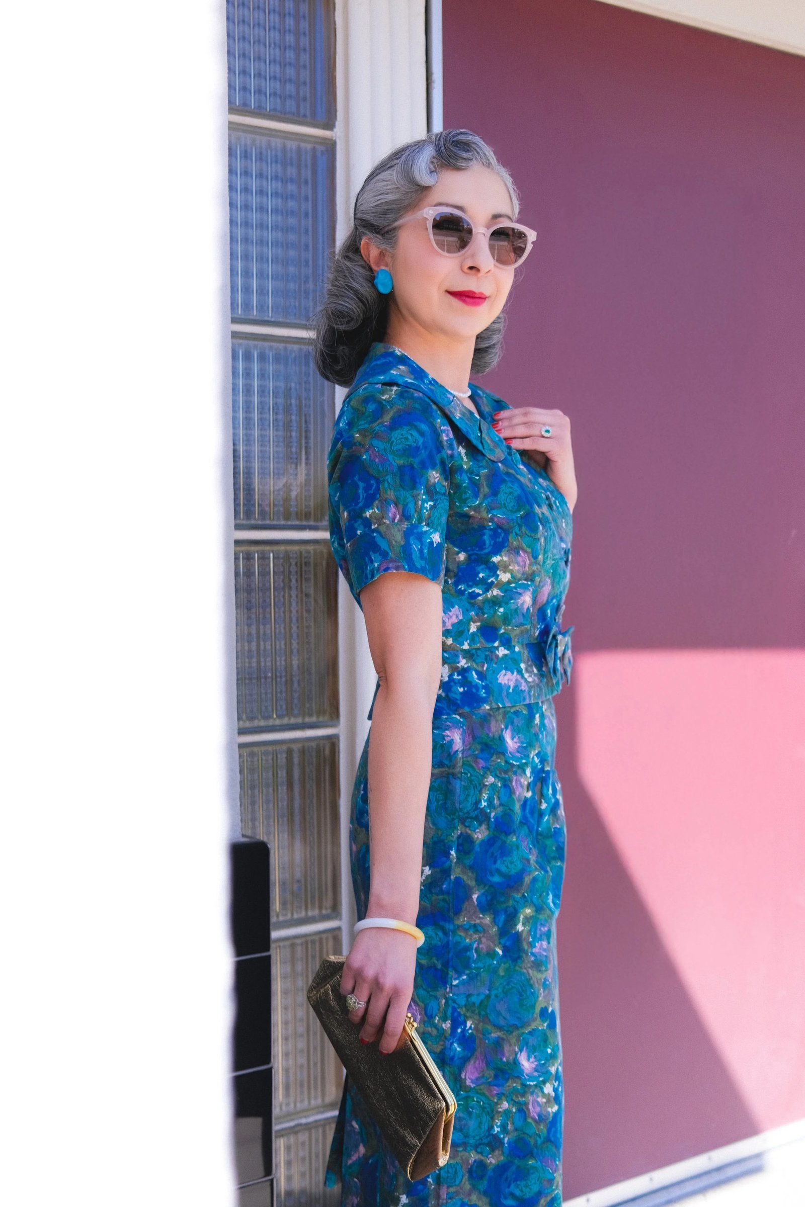 A woman in a vintage floral dress and retro sunglasses poses beside a building, holding a clutch purse.