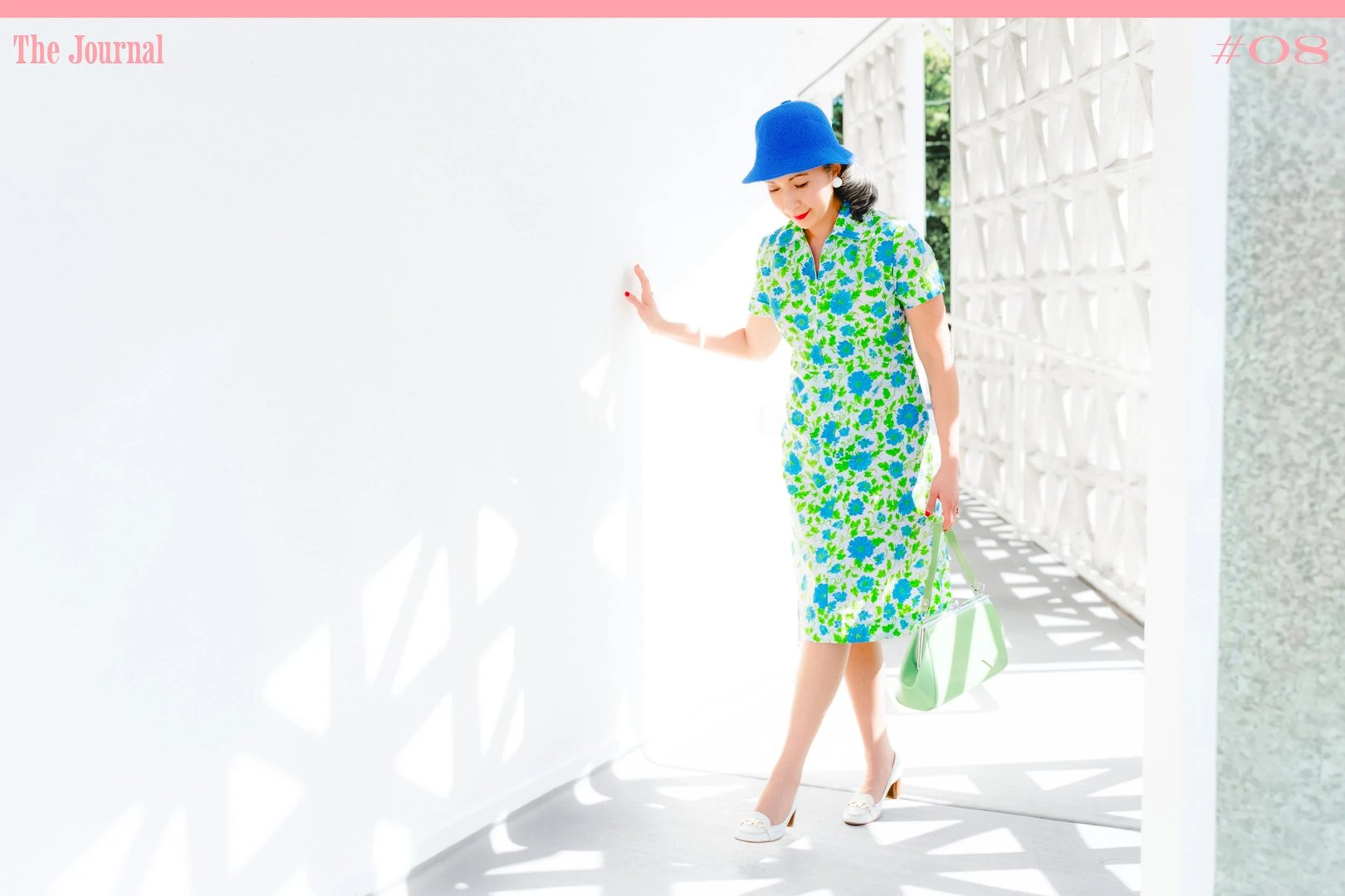 A woman in a floral print dress and blue hat poses against a bright white wall.