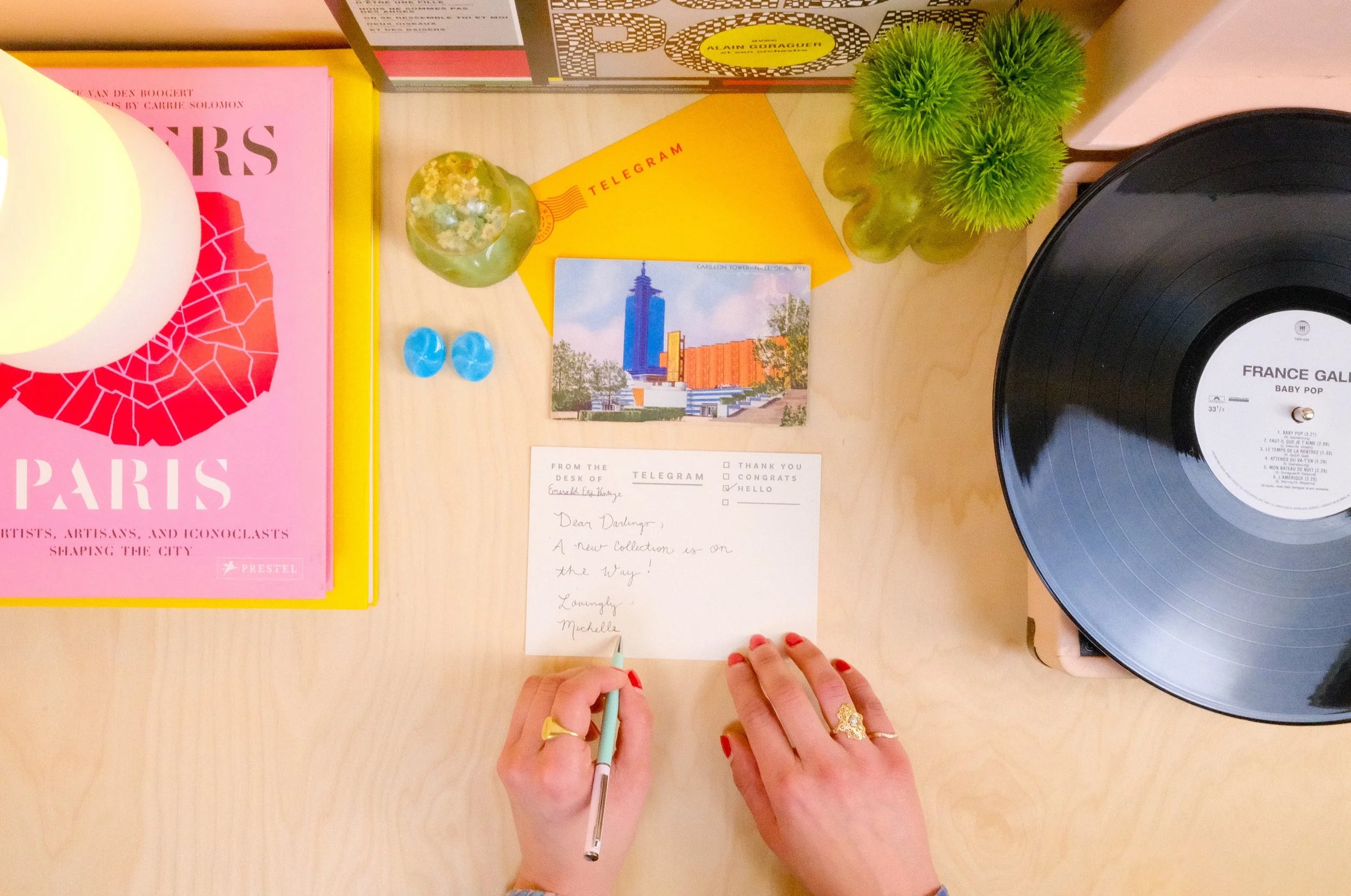A person writes a postcard on a wooden desk surrounded by books, a vinyl record, decorative plants, and stationery items.