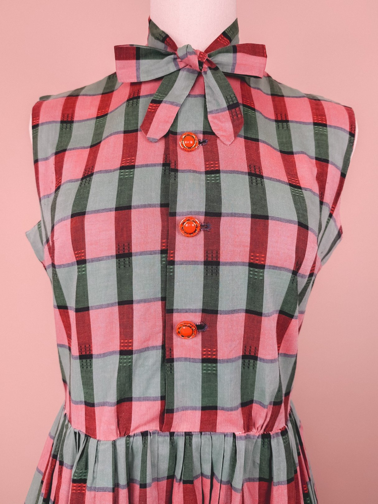 Red and green plaid dress featuring a high collar and bow tie, with three orange buttons down the front.