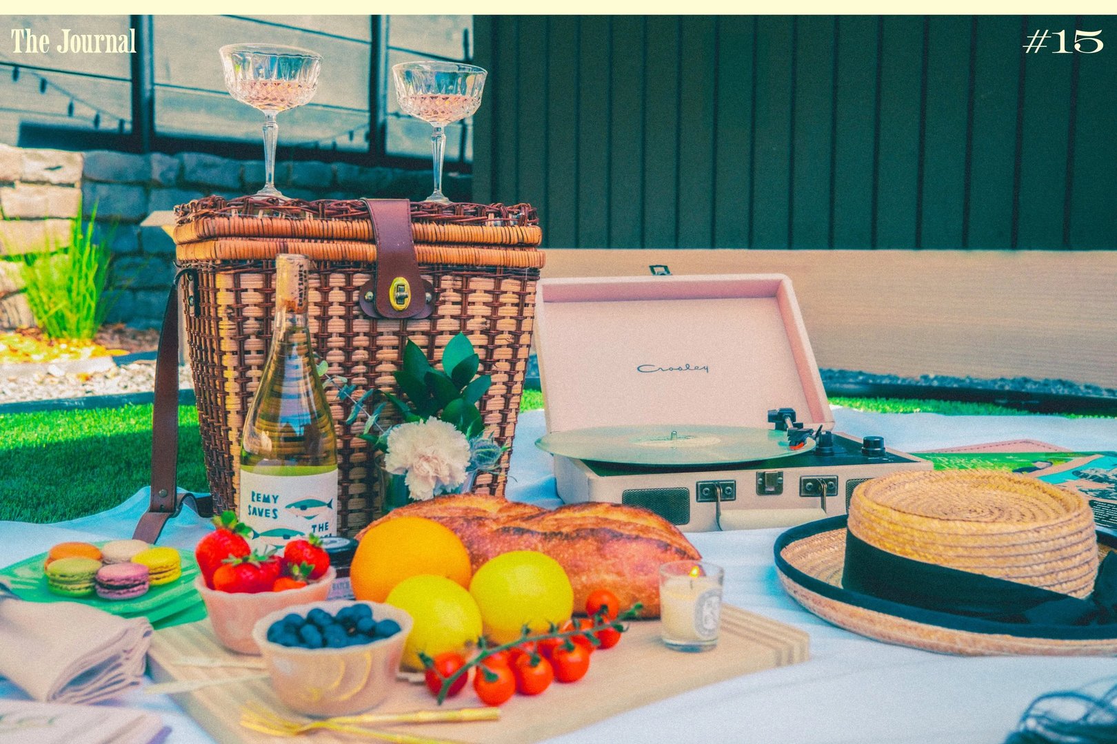 A picnic setup featuring a wicker basket, wine and a record player, with an assortment of fruits, pastries, and two glasses on a blanket.