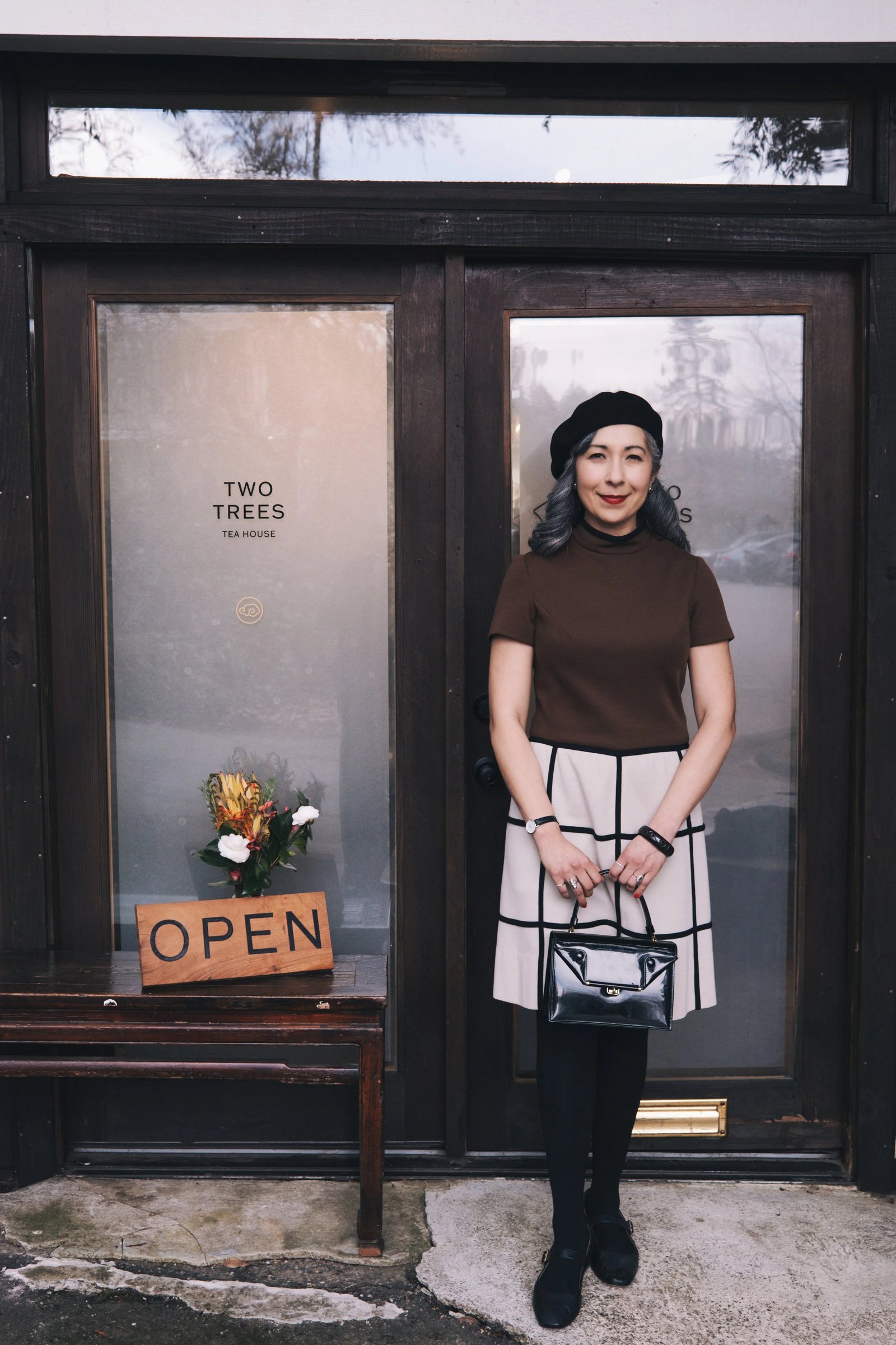 A woman in a brown top and plaid skirt stands outside a shop with an "OPEN" sign and a flower arrangement.