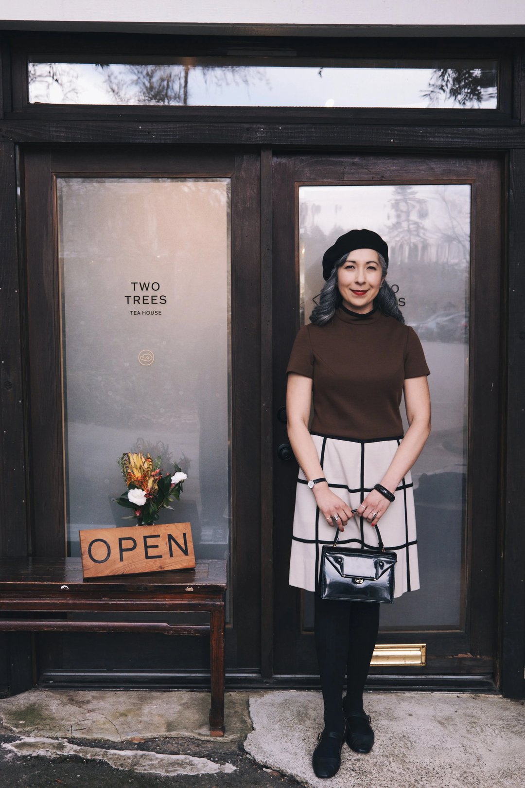 A woman in a brown top and plaid skirt stands outside a shop with an "OPEN" sign and a flower arrangement.