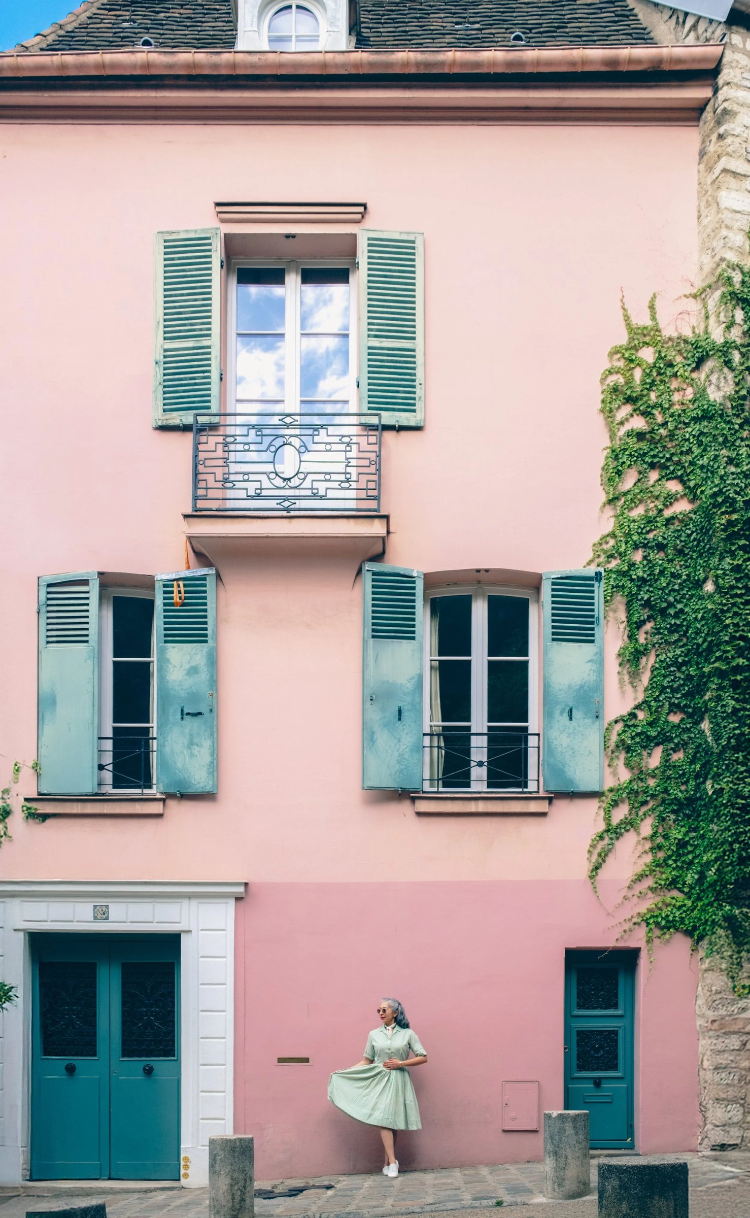 A pastel pink building with teal shutters and a woman in a light dress standing outside.
