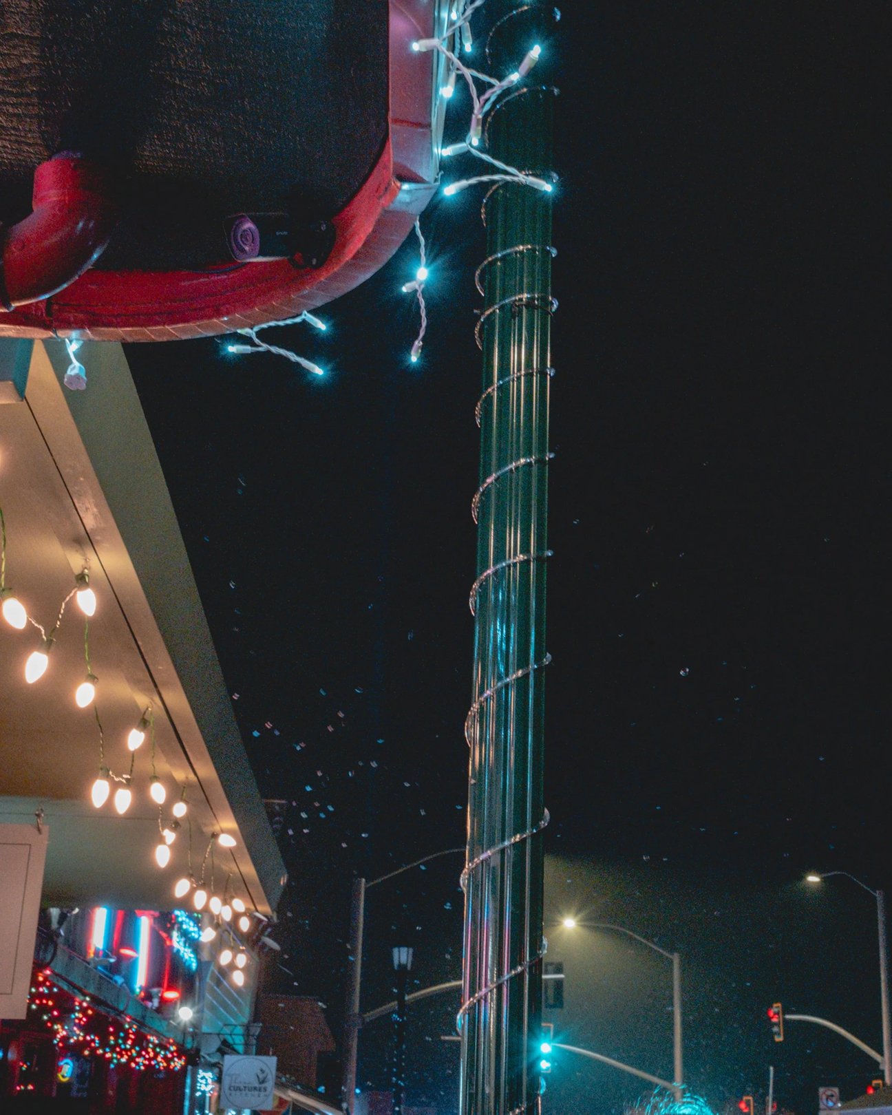 A city street at night featuring a lit pole adorned with blue decorative lights and blurred festive decorations in the background.