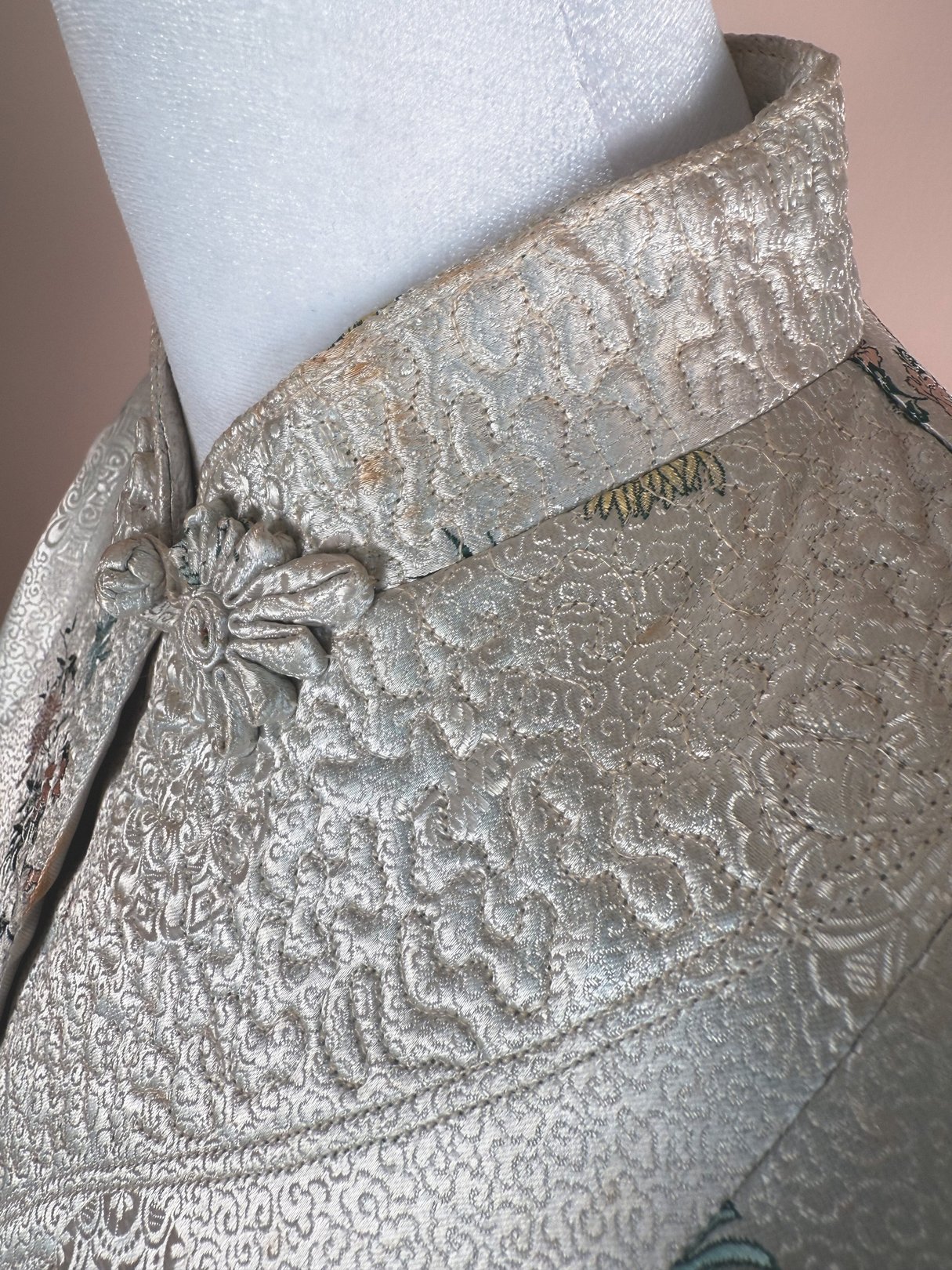 A close-up of a silver 1970s women's cheongsam coat featuring intricate embroidered patterns and a high collar with a decorative fastener.