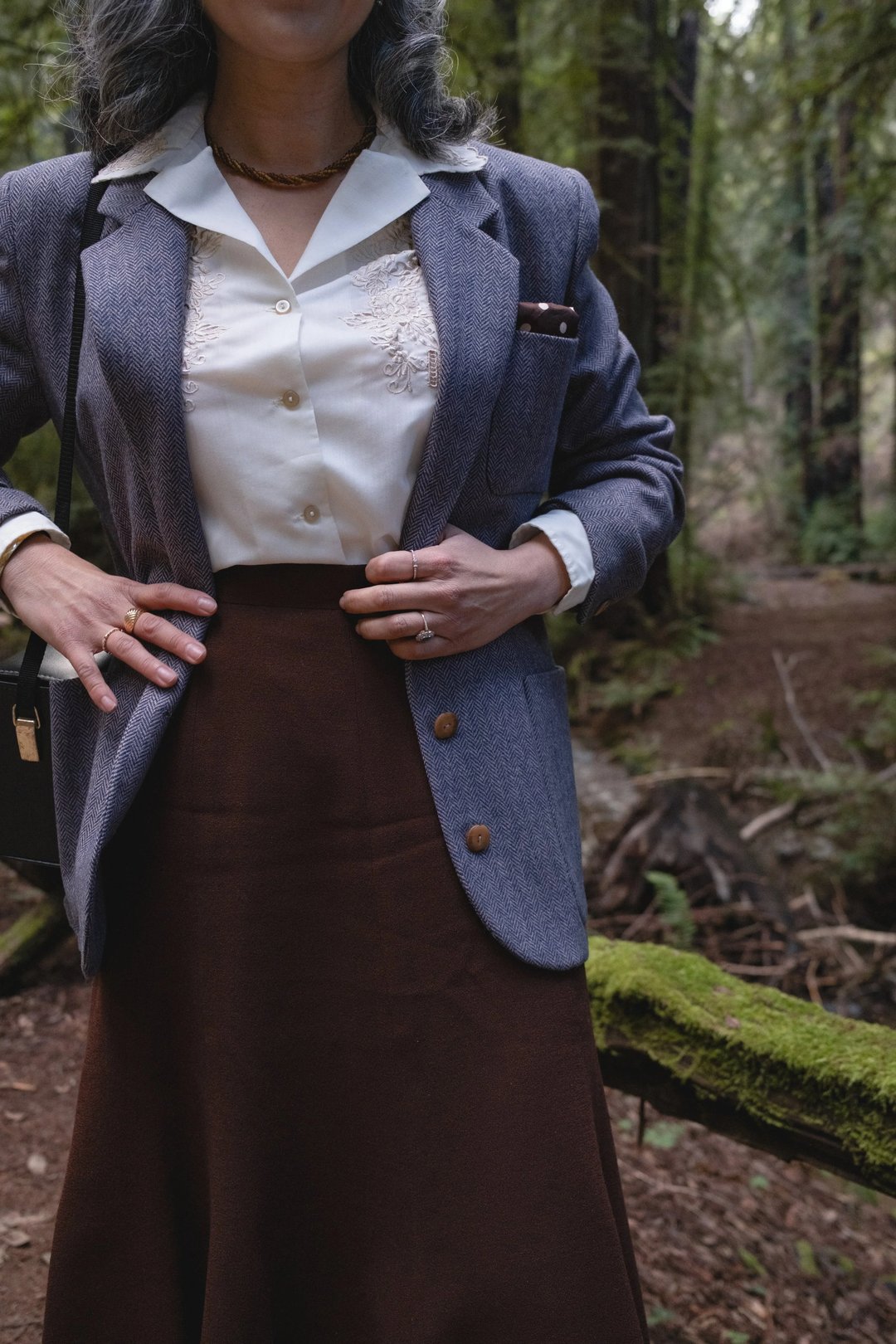 A person wearing a blue blazer over a white blouse and brown skirt stands confidently in a forest setting.