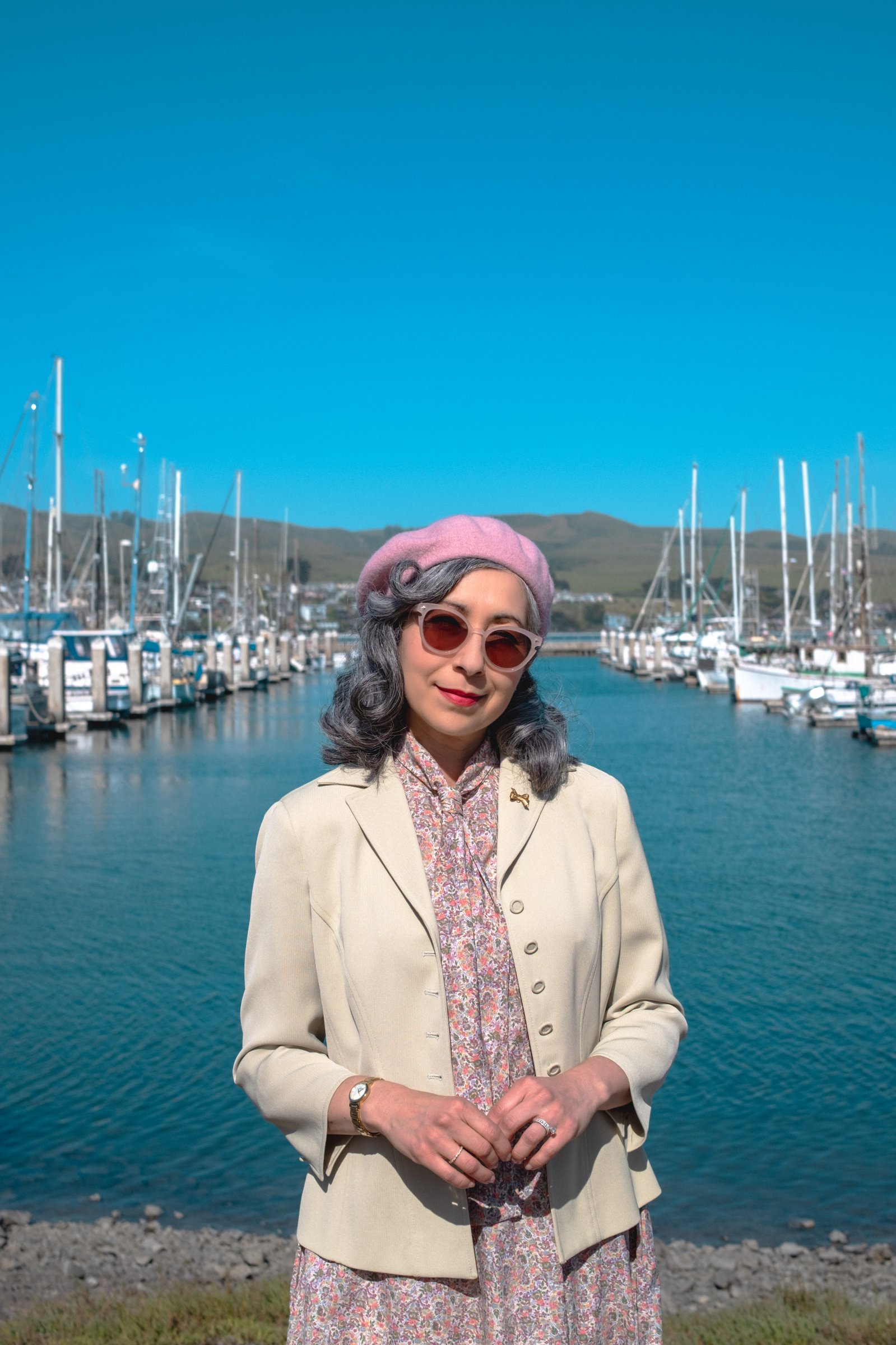 The Road to Bodega Bay gallery image: A woman in a pastel beret and sunglasses stands near a marina with sailboats and clear blue skies in the background.