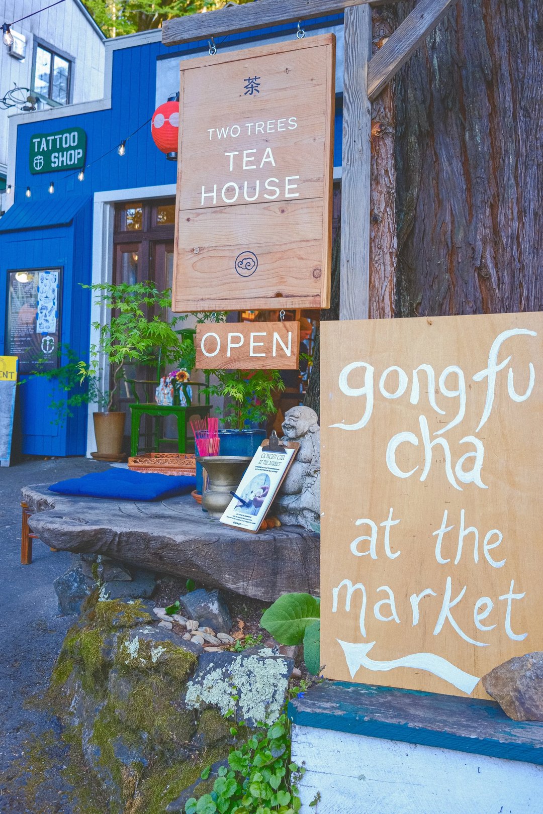 A wooden sign for a tea house with the text "gongfu cha at the market" and decorations in a vibrant outdoor setting.