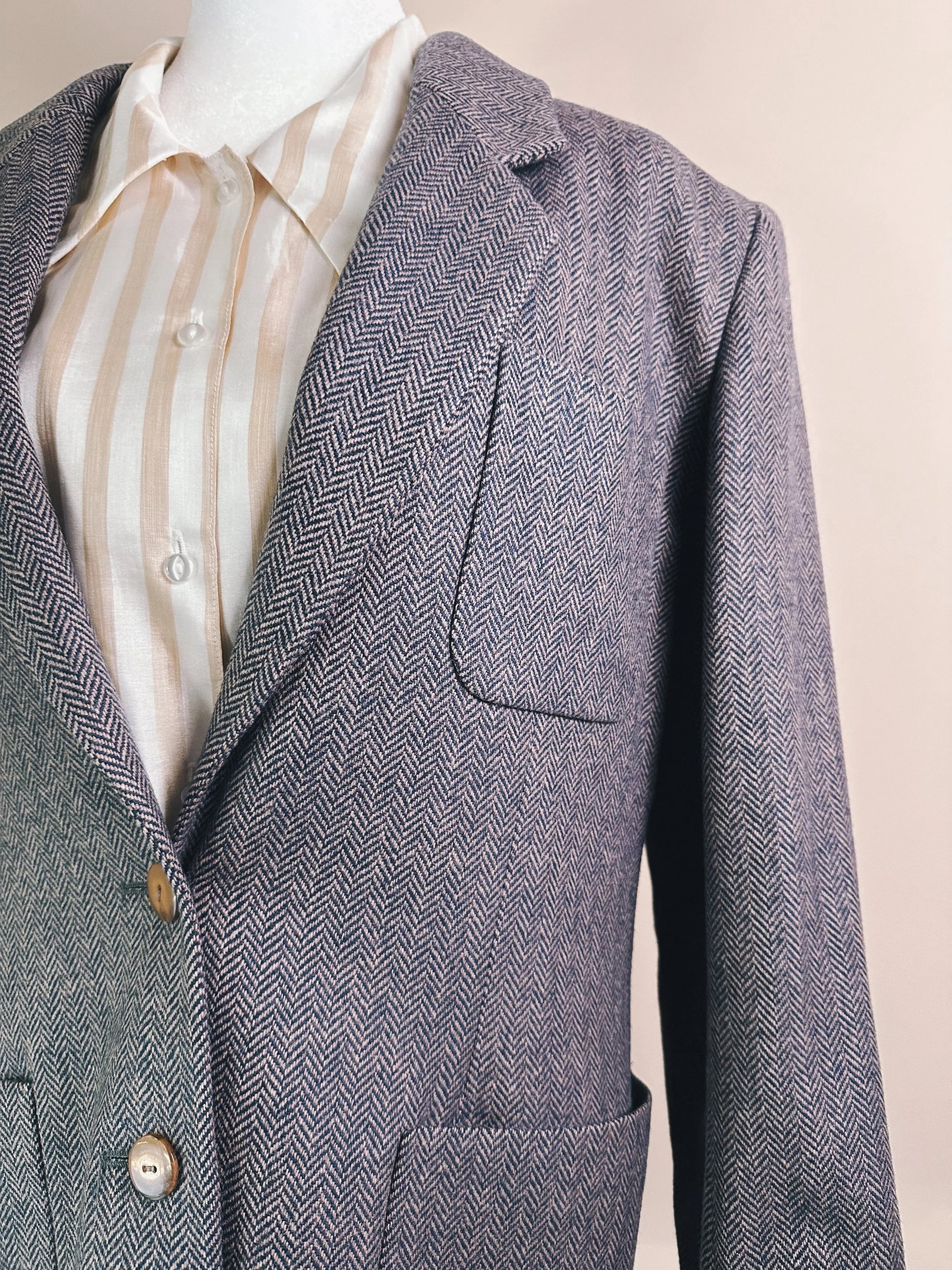 A gray herringbone blazer displayed on a mannequin, featuring a single chest pocket and two front pockets, layered over a light striped button-down shirt.
