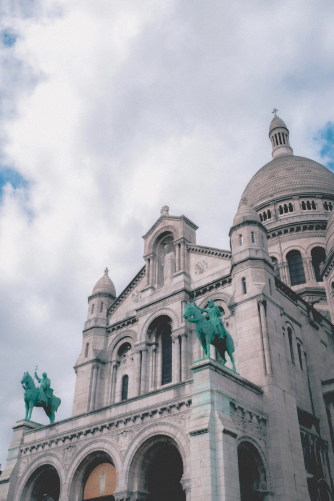 The Sacré-Cœur Basilica in Paris, featuring two green horse statues at the entrance, against a backdrop of cloudy skies.