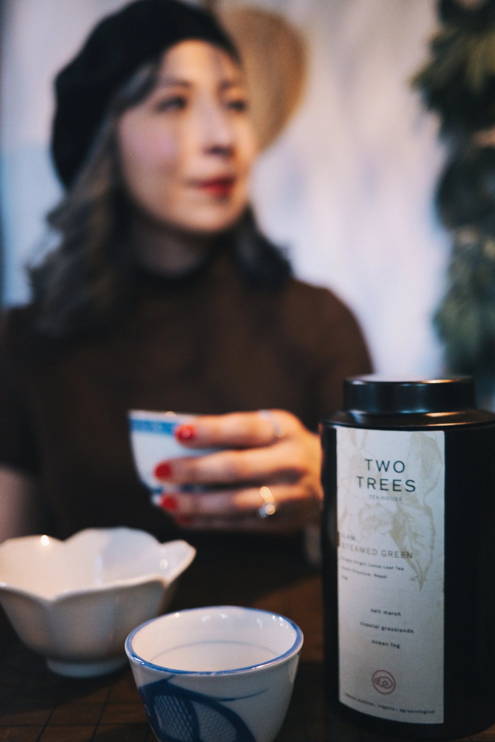 A woman holds a tea cup while sitting at a table with a canister of Two Trees tea and a small dish in the foreground.