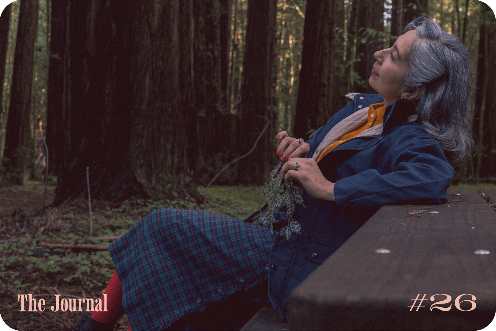 A person wearing a blue jacket and plaid skirt sits on a bench in a forest, holding a small branch.