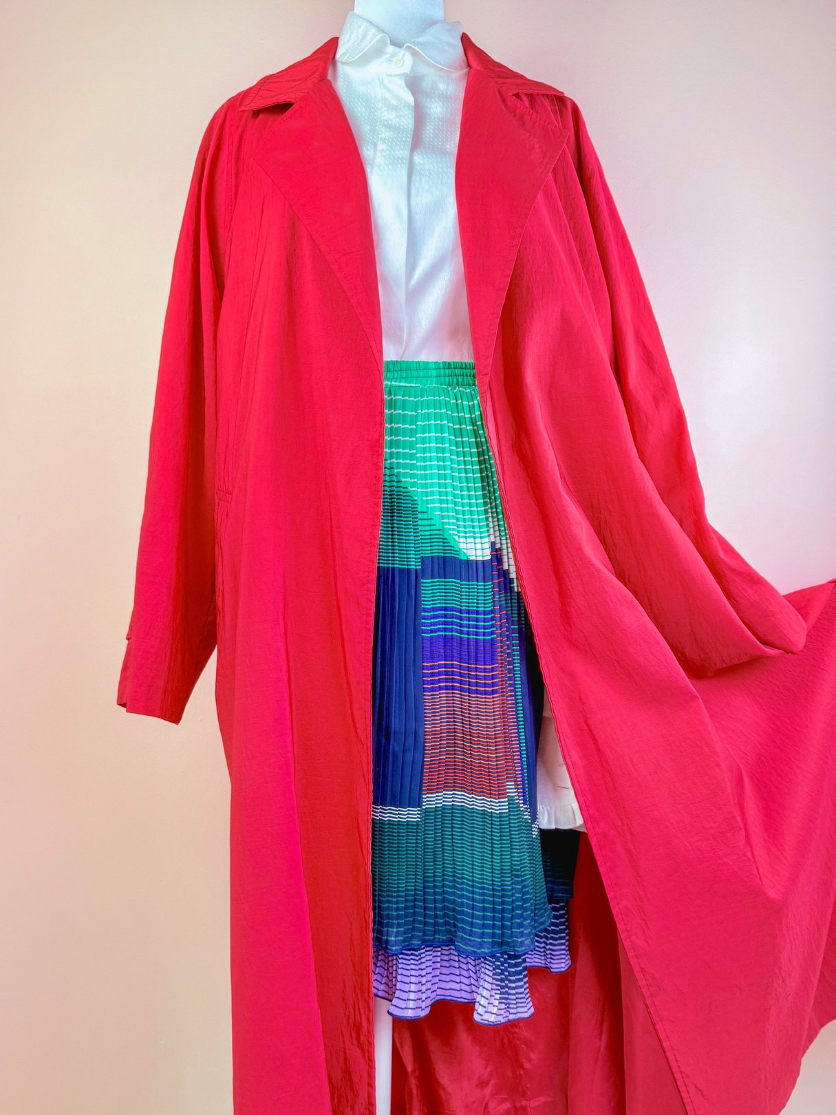 A vibrant red 1980s women's rain jacket displayed with a white blouse and colorful striped skirt.