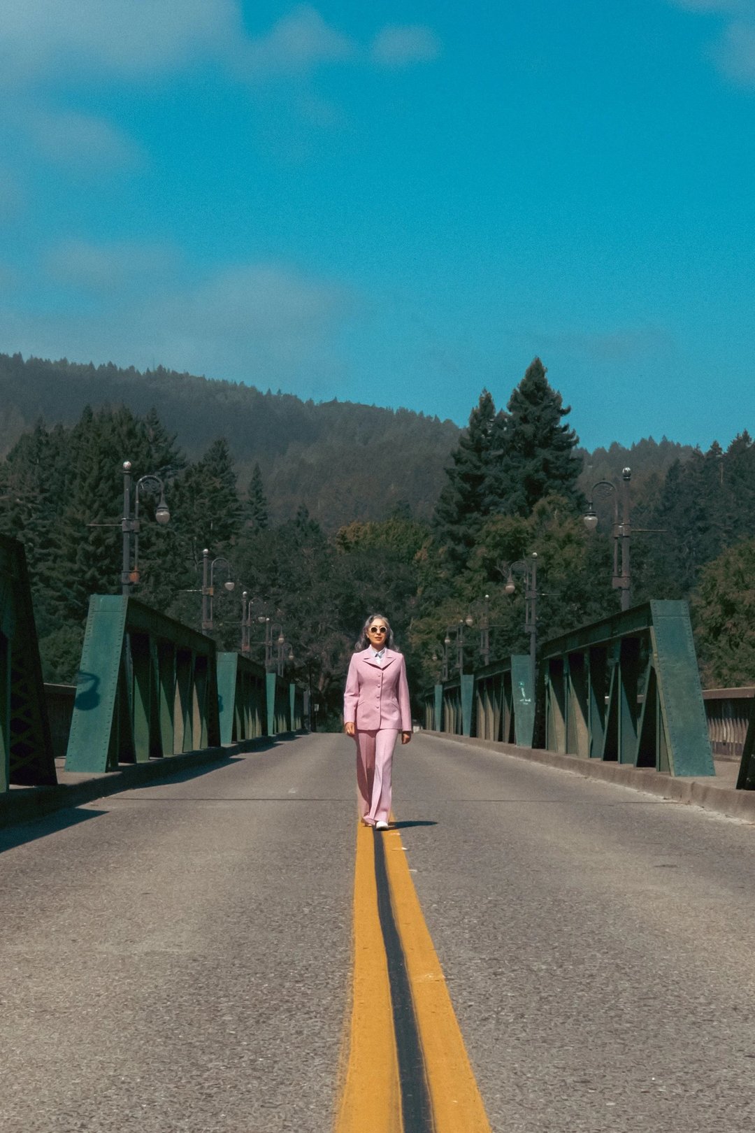 A person wearing a pink suit walks alone on a deserted bridge surrounded by trees and mountains under a blue sky.
