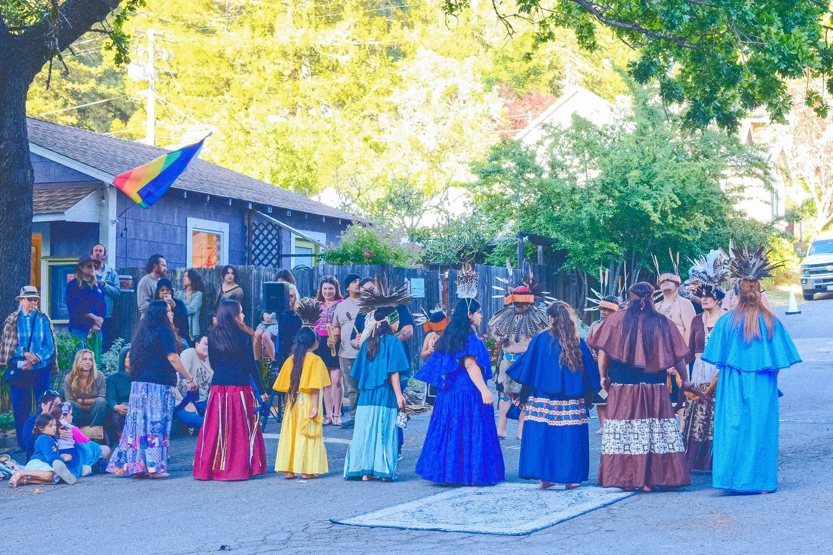 A diverse group of performers in traditional attire dances in a street surrounded by an audience, with trees and buildings in the background.