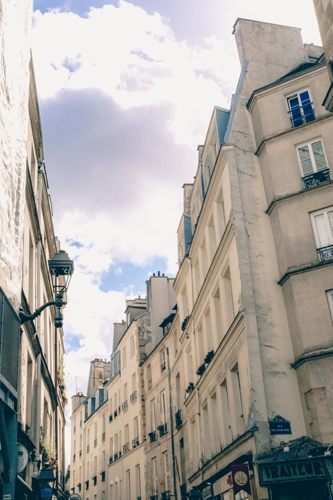 A narrow street in a European city featuring tall buildings and a vintage streetlamp under a partly cloudy sky.