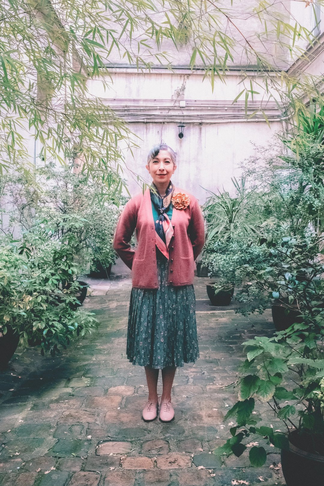 A woman stands in a garden surrounded by potted plants, wearing a pink cardigan, floral dress, and styled hair with a large flower accessory.