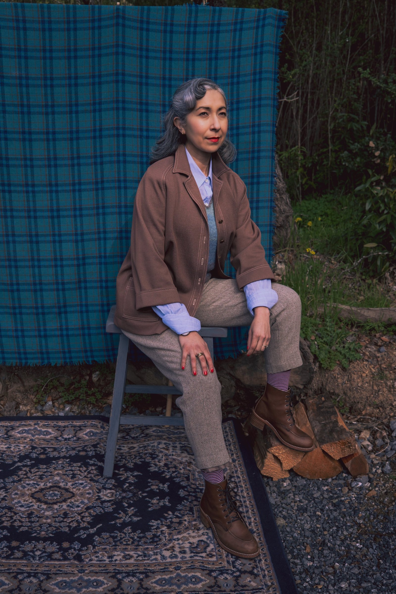 A woman with gray hair styled in waves sits on a chair in front of a plaid backdrop, wearing a brown cardigan, gray vest, and tailored trousers, complemented by brown boots.