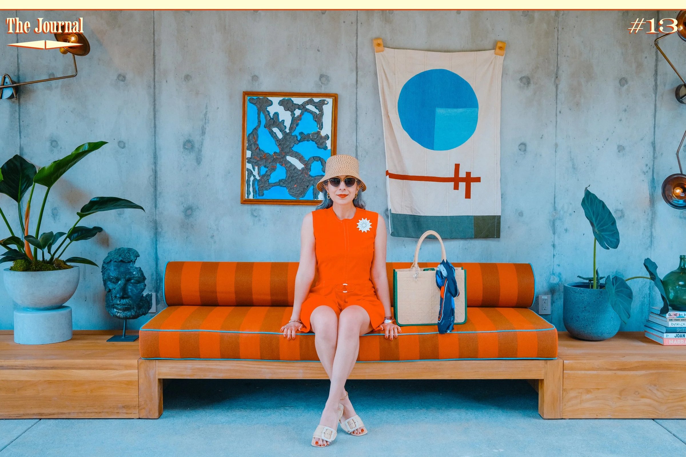 A woman in a red dress and sun hat sits on an orange-striped couch surrounded by greenery and modern decor.