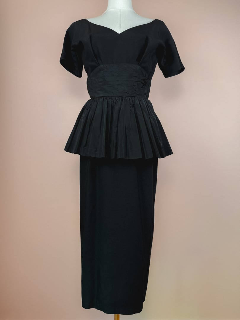 Black 1950s-style dress featuring short sleeves, a fitted bodice, and a pleated peplum waist.