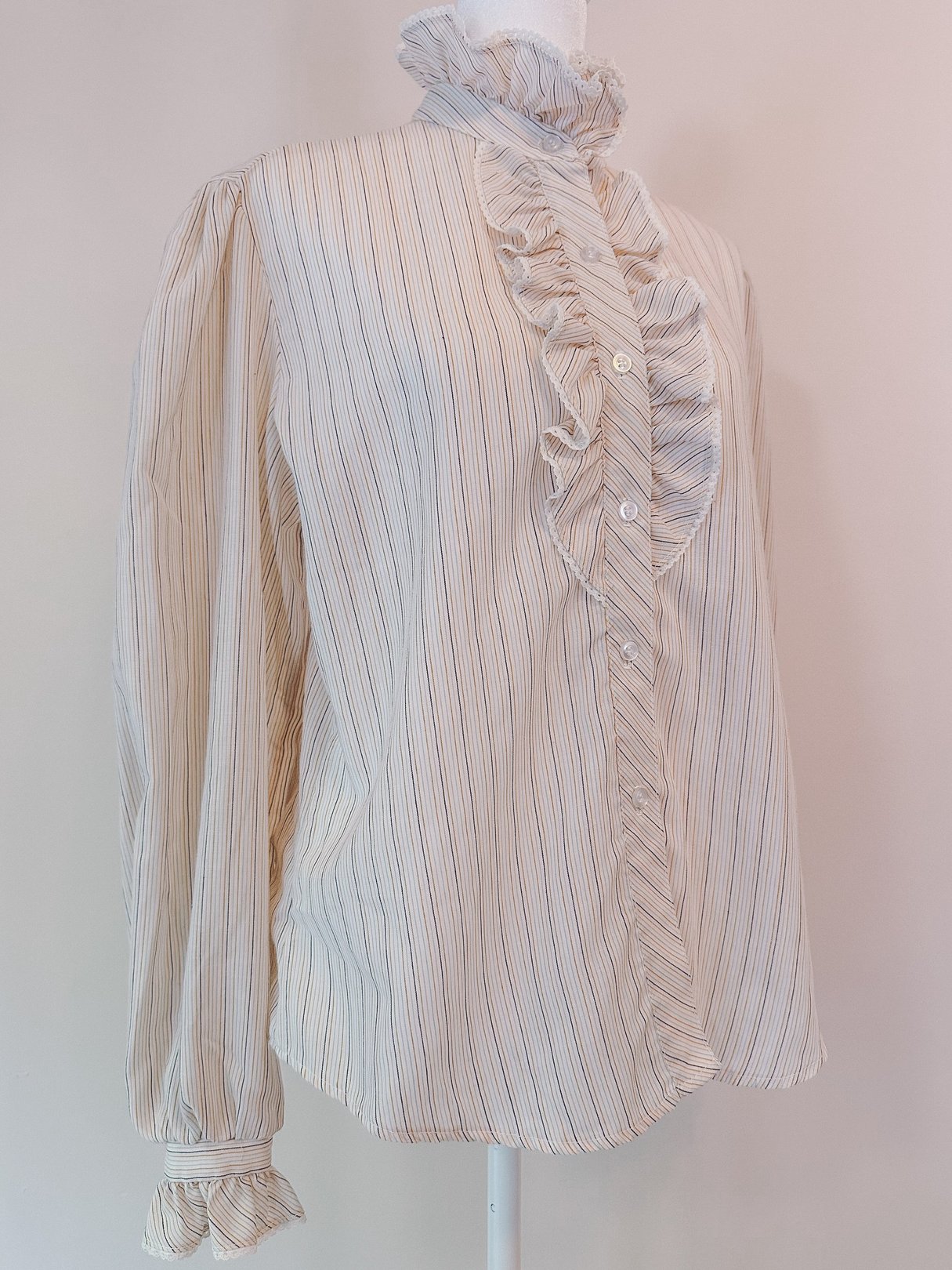 Cream blouse with ruffled neck and long sleeves, featuring intricate pink, orange, and blue stripes, styled for women and inspired by the 1970s.