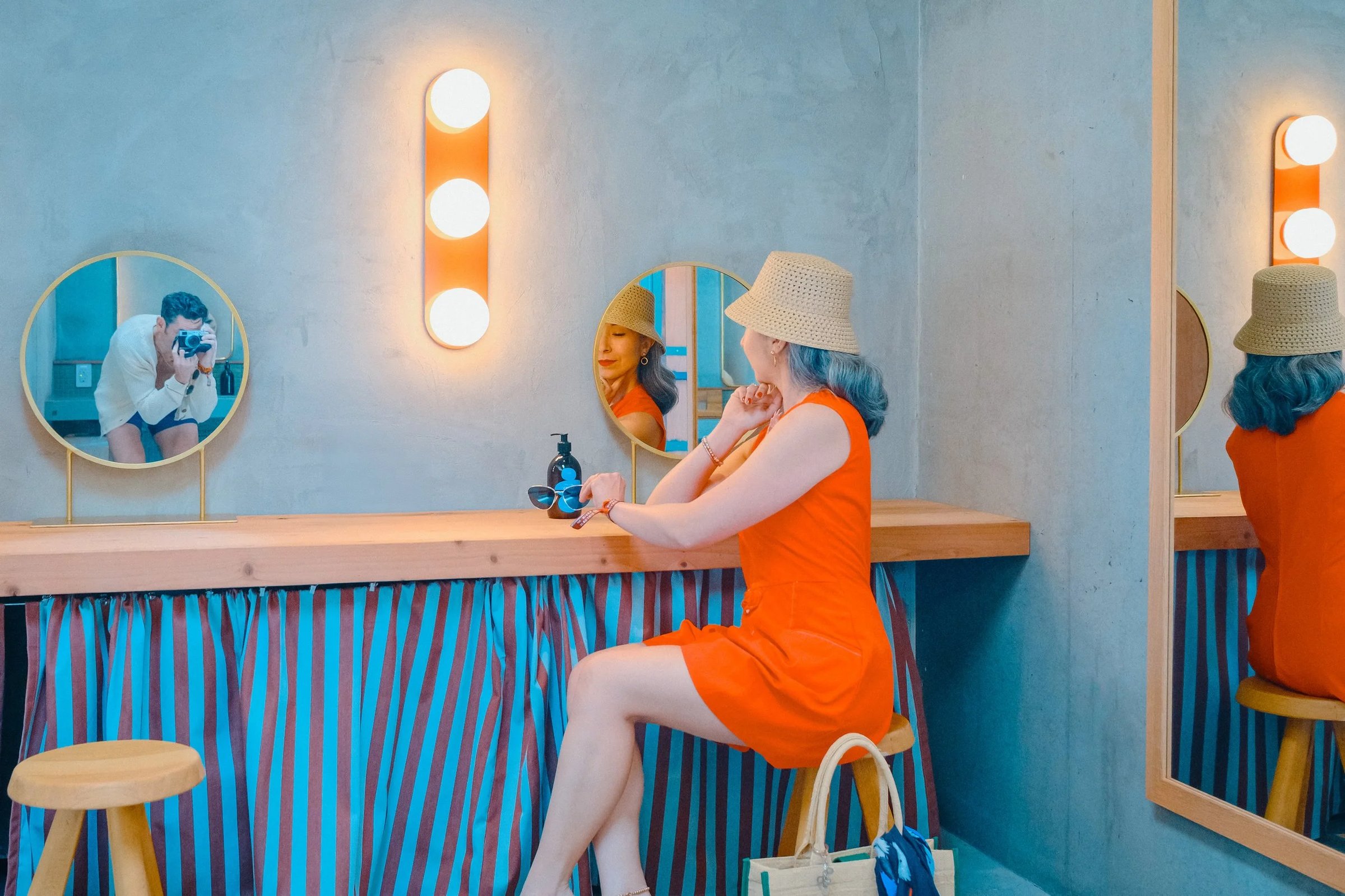 A woman in an orange dress and straw hat sits at a counter with a retro-styled mirror, while a person with a camera is reflected behind her.