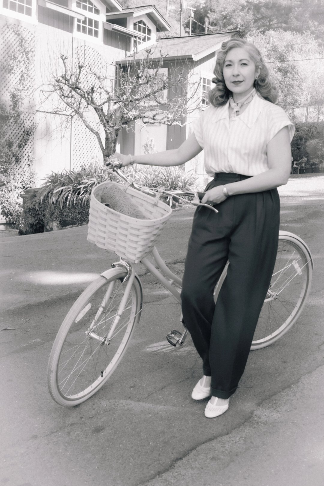 A woman in a striped blouse and high-waisted trousers poses next to a vintage bicycle with a wicker basket.