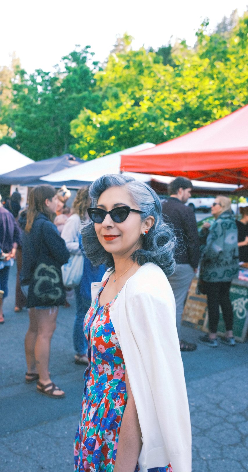 A woman with silver hair and sunglasses poses in a colorful floral dress at a lively outdoor market surrounded by people.