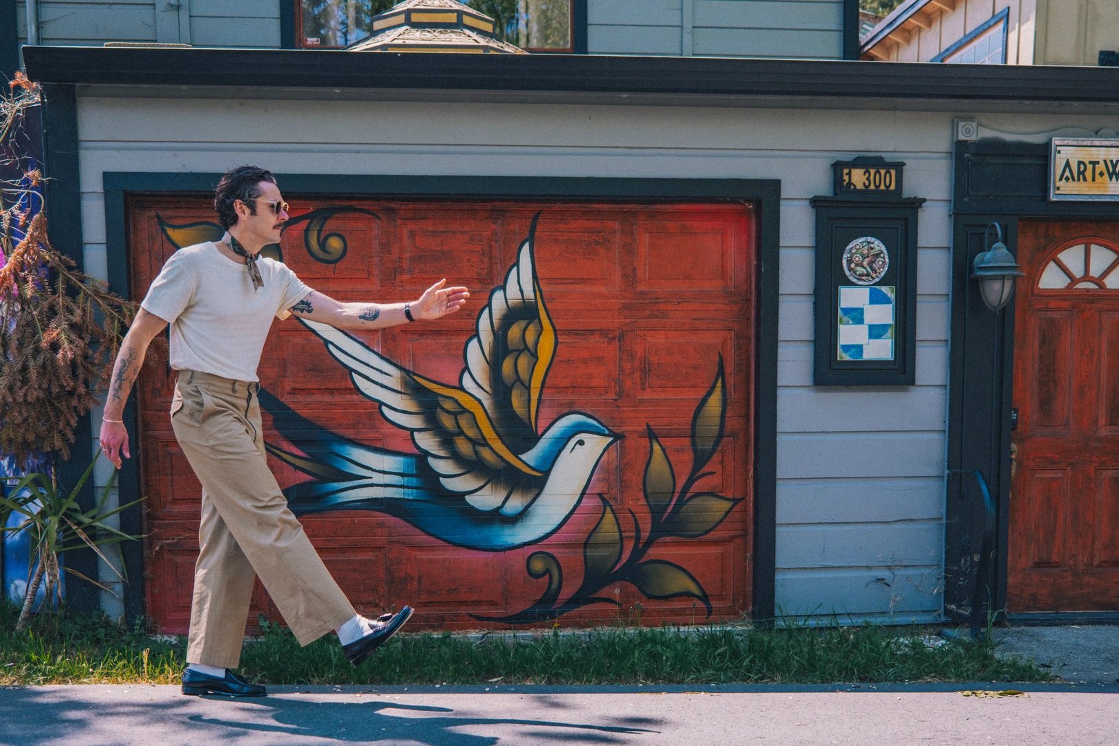 A man in a cream sweater and beige pants walks past a colorful mural of a bird on a garage door.