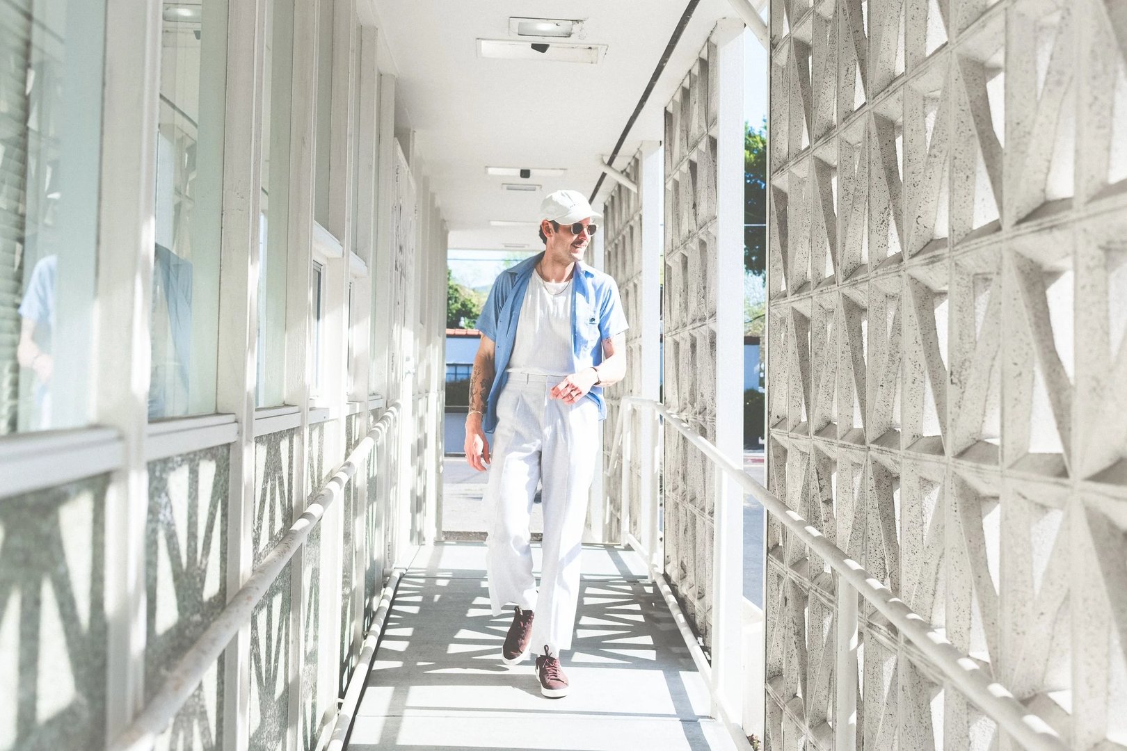 A man wearing a light blue shirt, white trousers, and a white cap walks through a sunlit hallway with patterned walls.