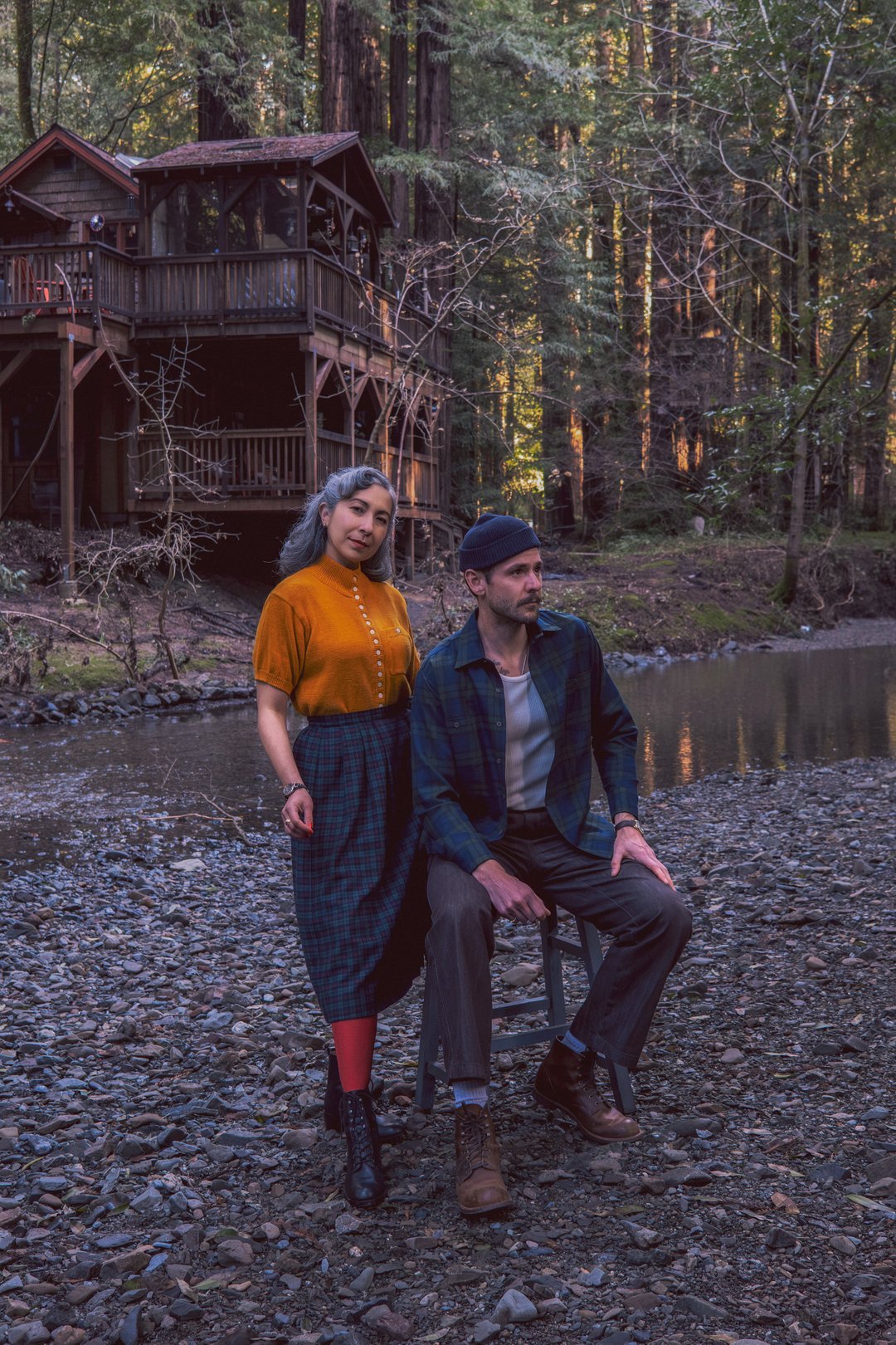 A man and woman pose together by a tranquil stream in a wooded area, surrounded by trees and a cabin in the background.