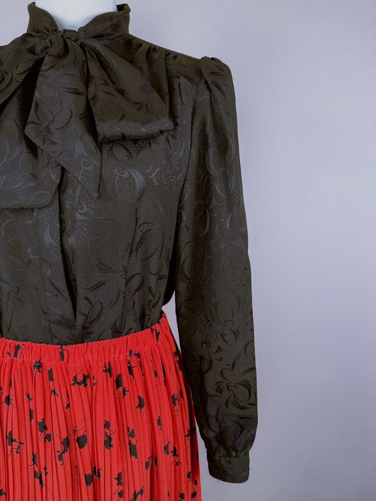 Red pleated skirt with a floral pattern, paired with a black blouse featuring a high collar and bow detail, representing women's fashion from the 1970s.