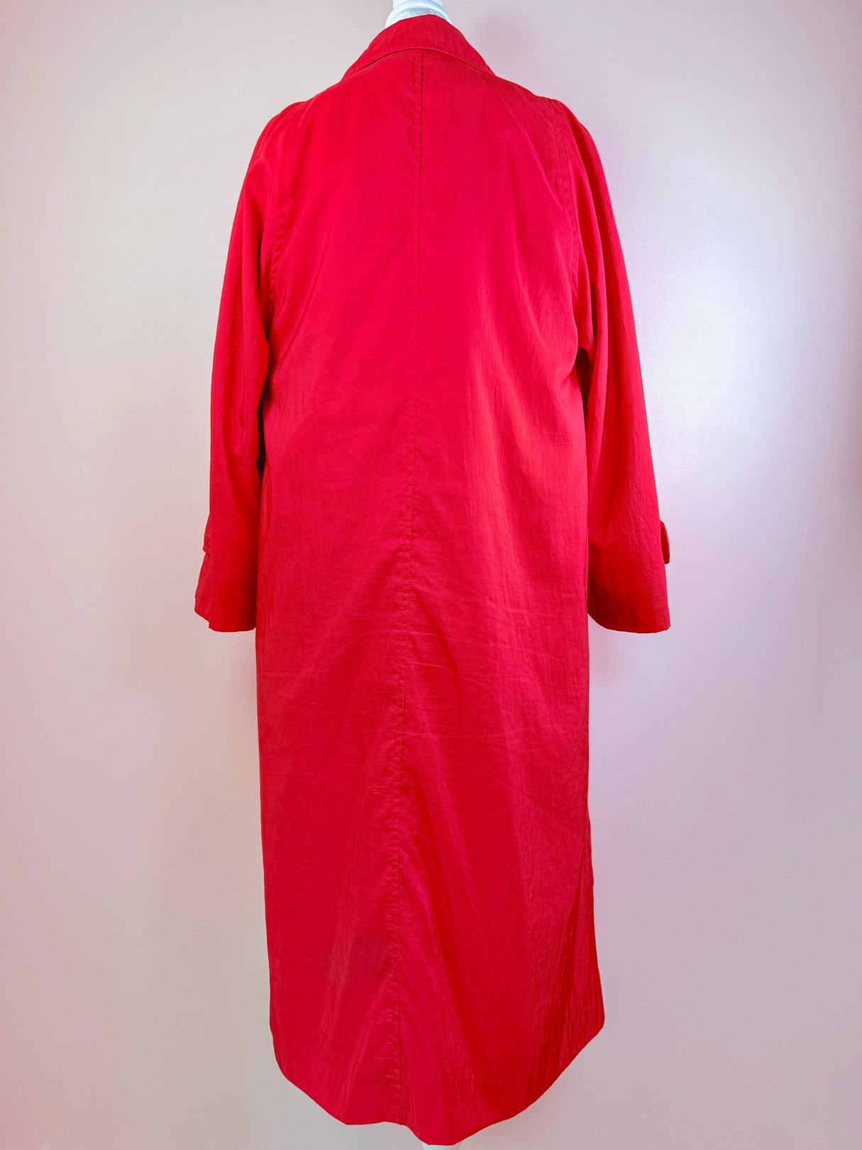 A red 1980s women's rain jacket with a long silhouette and back pleat displayed against a neutral background.