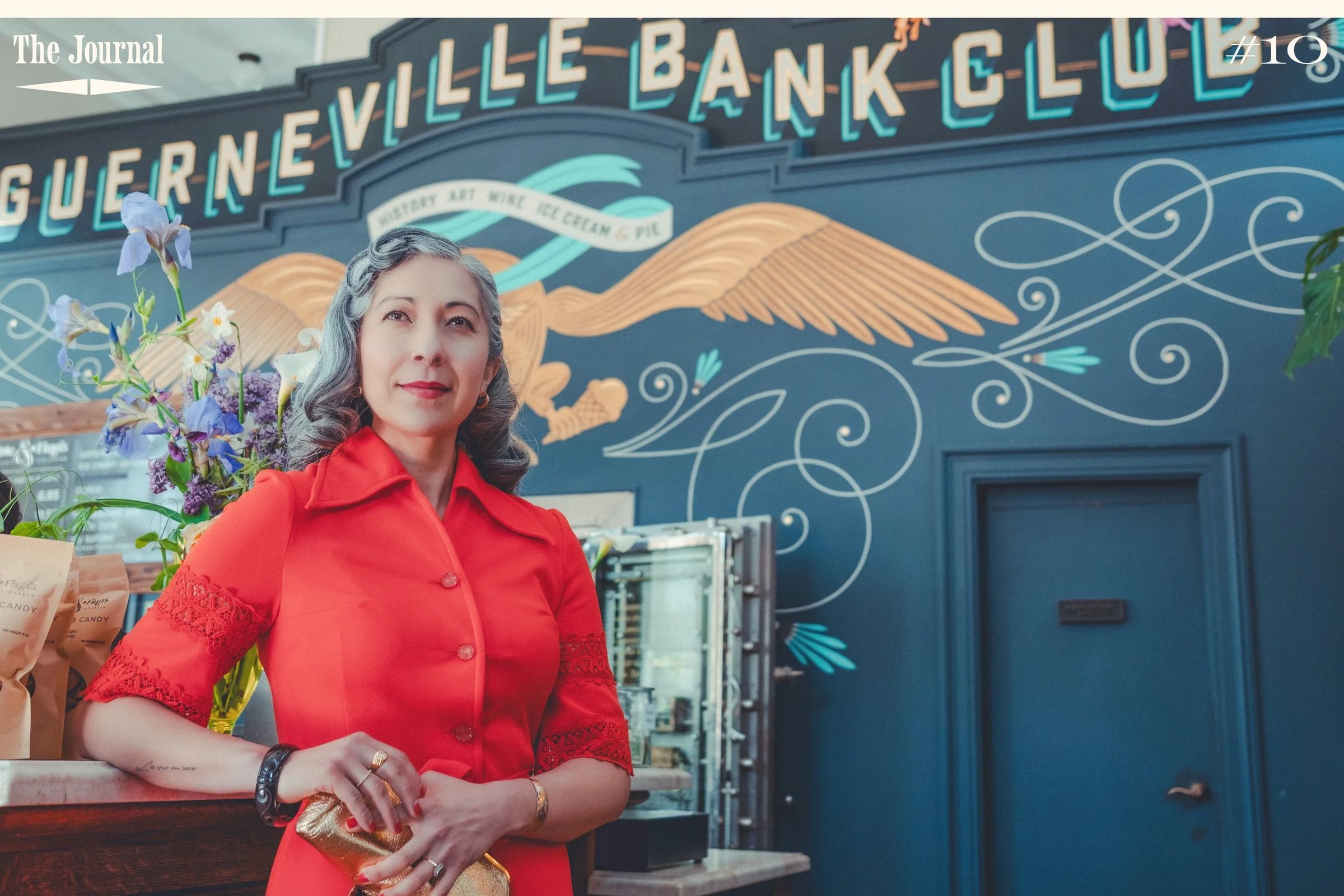 A woman in a red dress stands in front of a decorative wall featuring the Guerneville Bank Club logo.