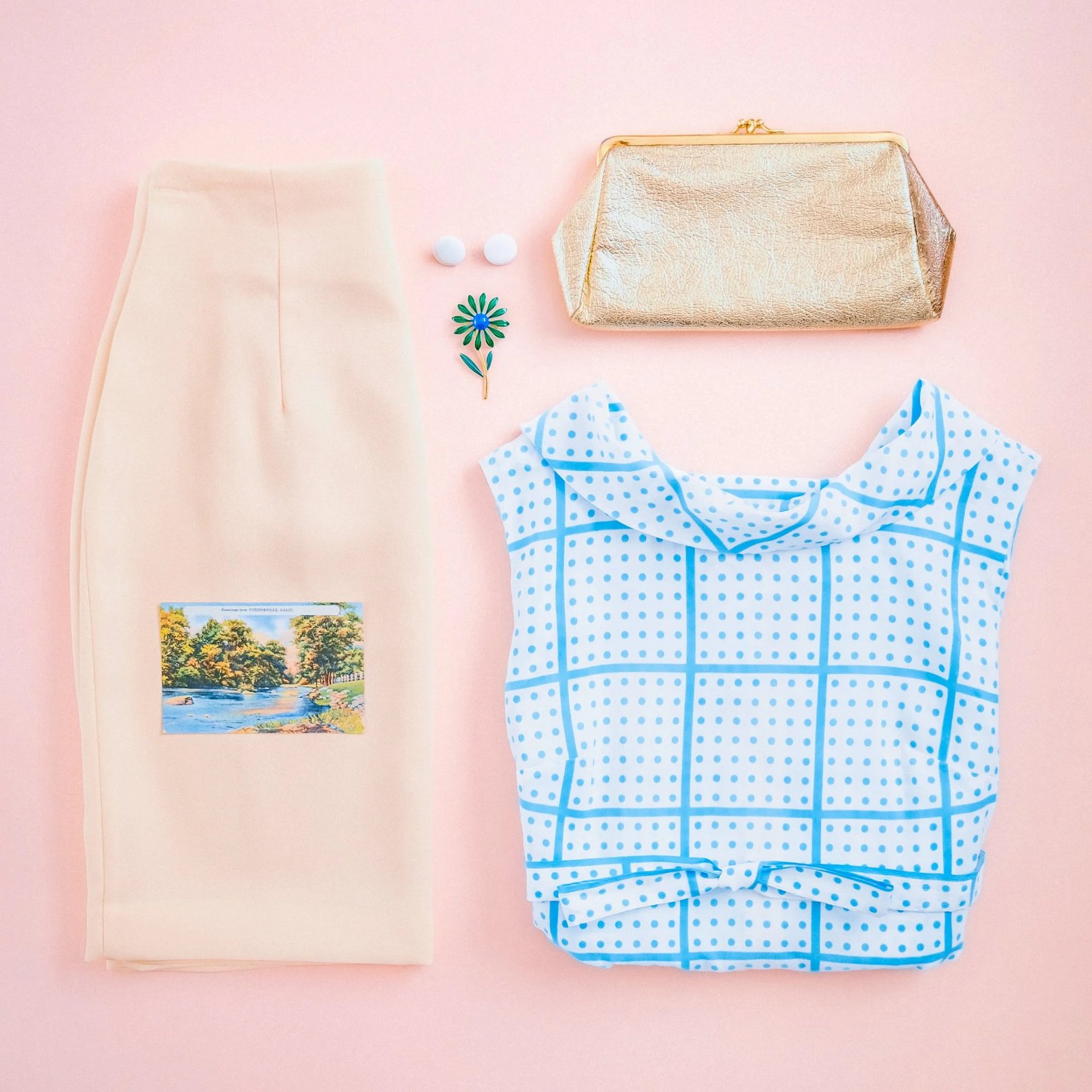 A flat lay arrangement featuring a cream skirt with a graphic design, a light blue and white checked shirt, a beige bag, and a green button on a pink background.